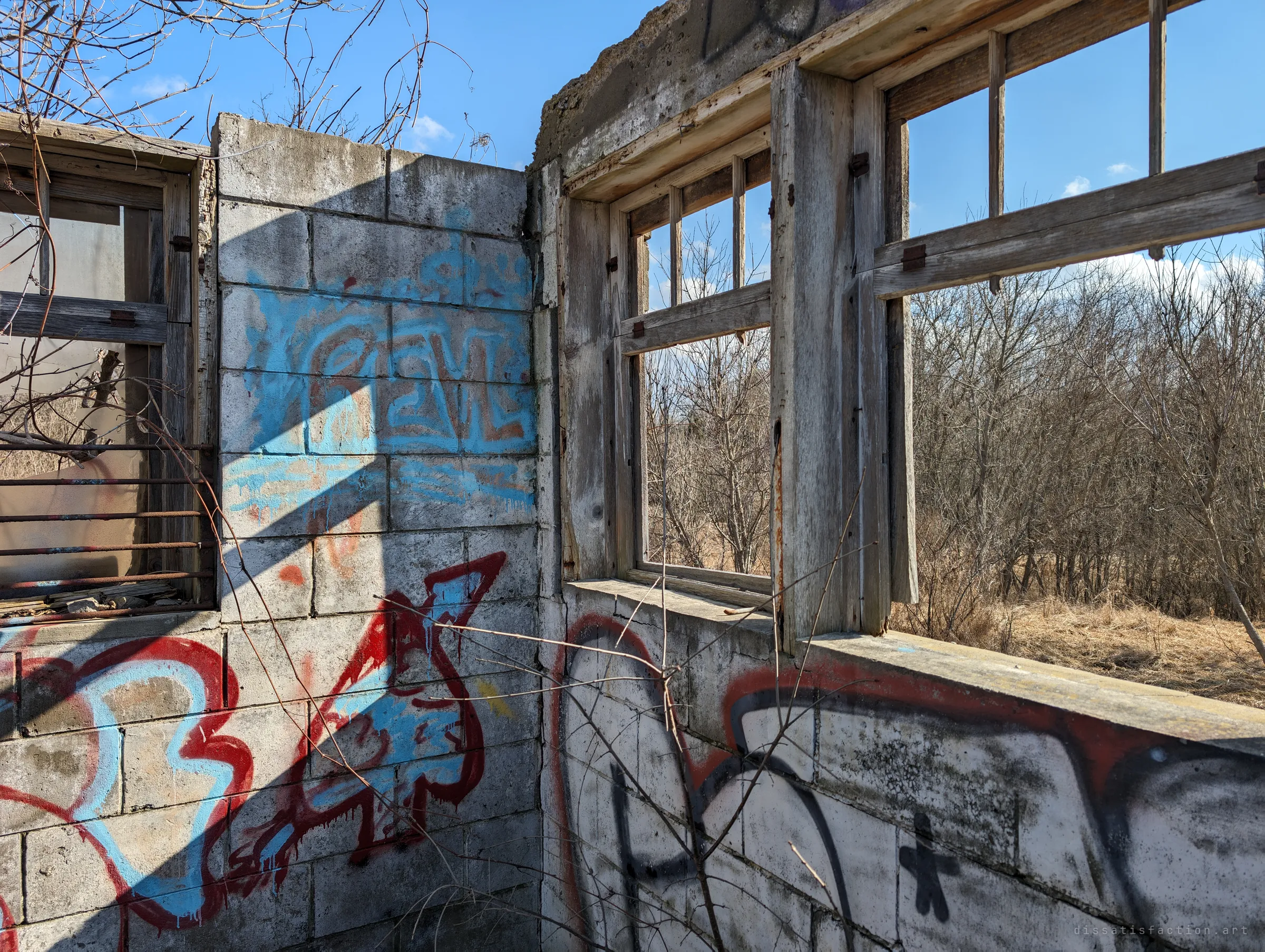 The inside corner of a roofless block building, its walls covered in red, blue and black graffiti, with empty wooden window frames looking out onto bare scrub. Hard shadows from the frames fall across the paint.