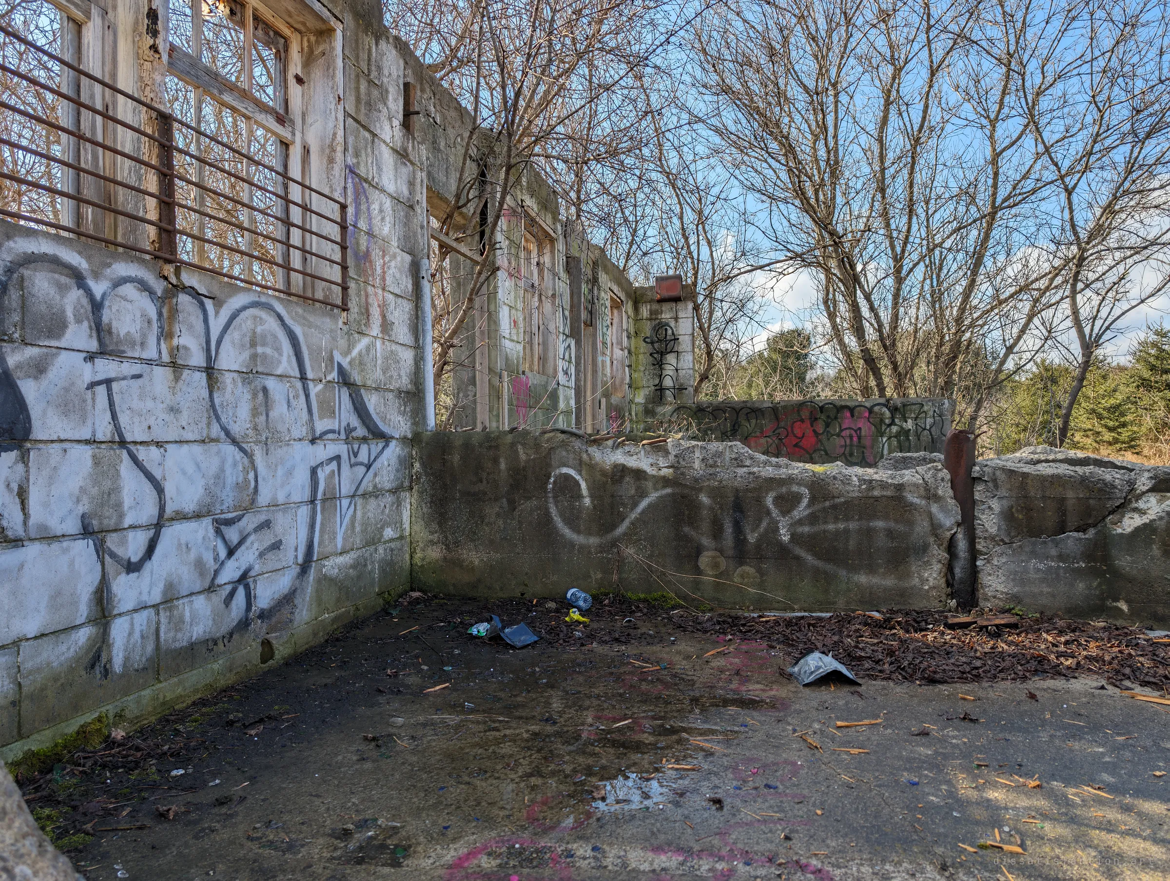 A graffitied concrete yard beside a barred-window block building, with broken low walls, puddles and scattered litter. The row of rooms stretches back toward the trees.