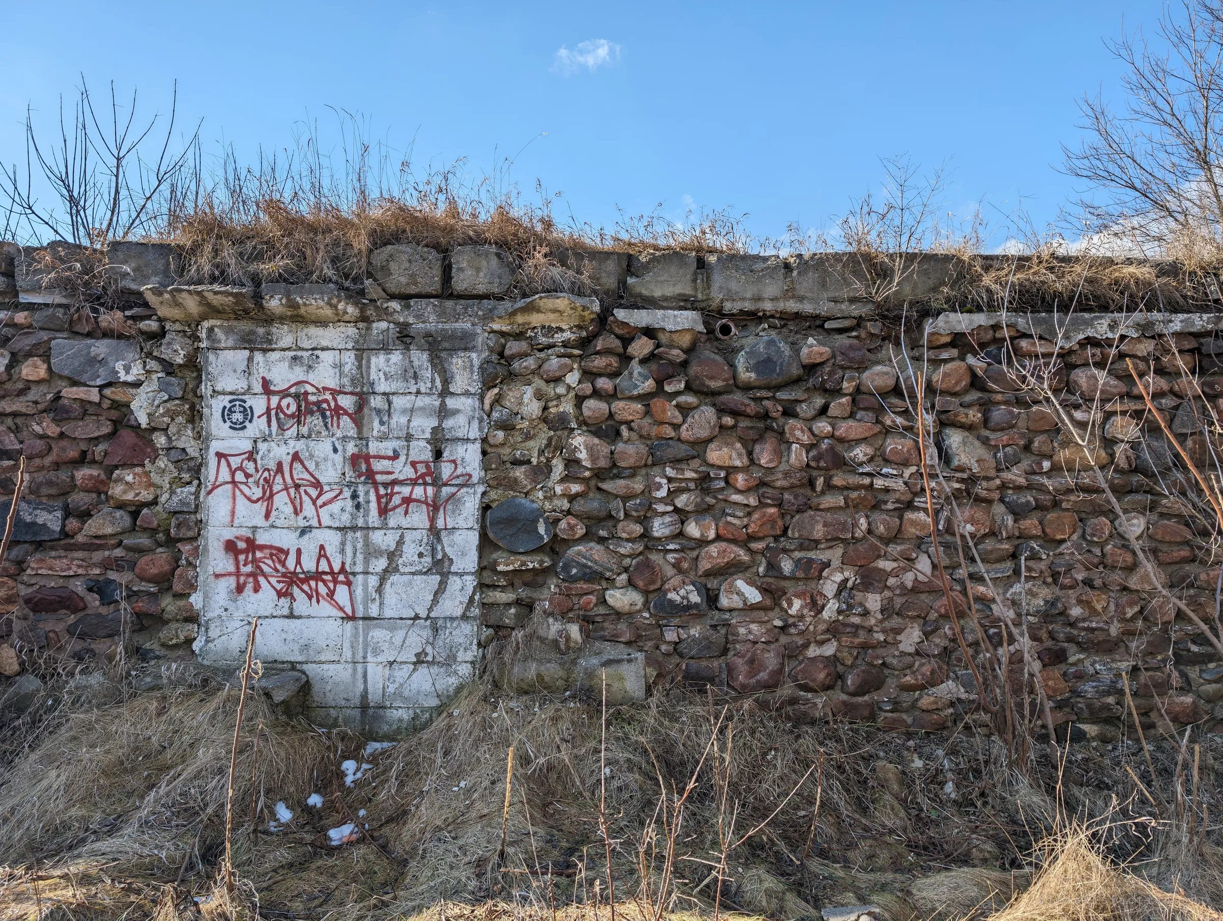 A fieldstone wall with a bricked-up doorway painted white and tagged in red, dry grass growing along its top. The mix of rounded river stones and block infill shows the building's patched history.