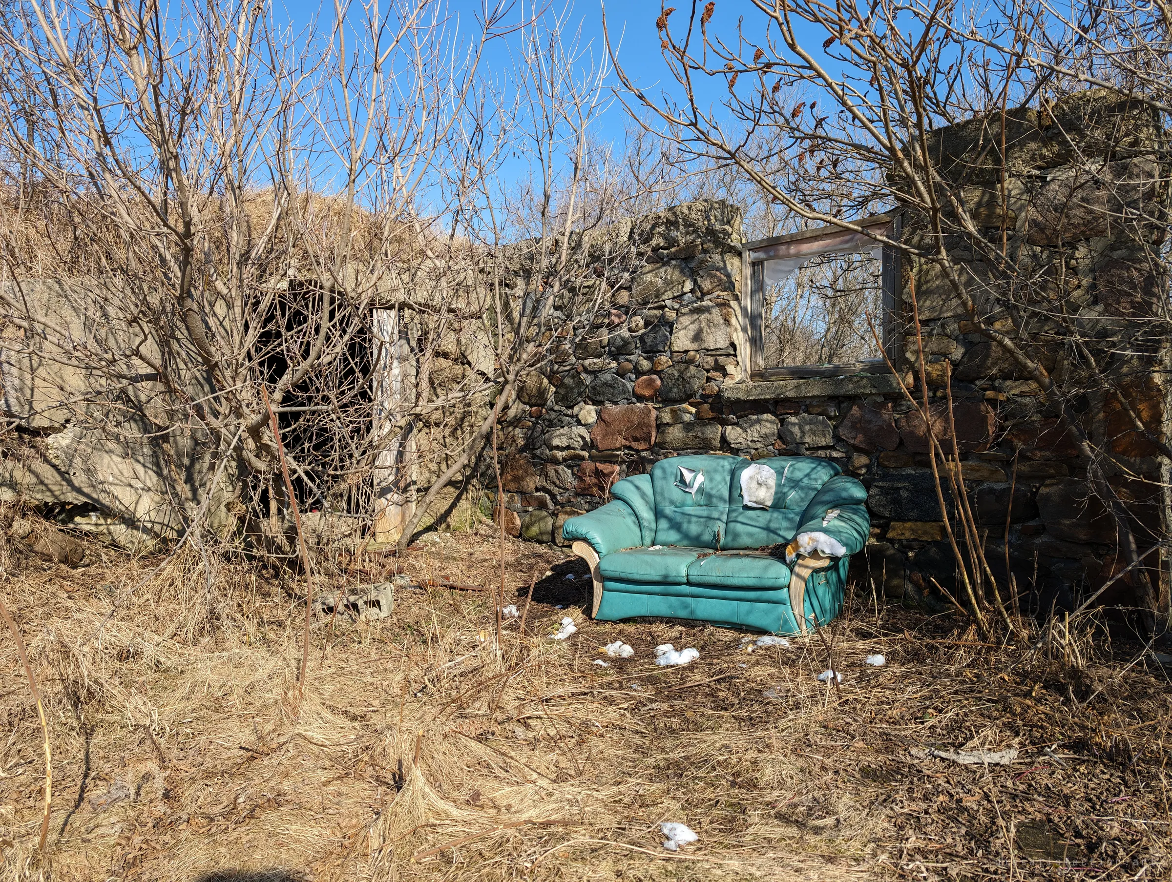 A torn teal leather loveseat sits in dry grass in front of a crumbling stone wall with an empty window frame. Someone has turned the ruin into an outdoor living room.