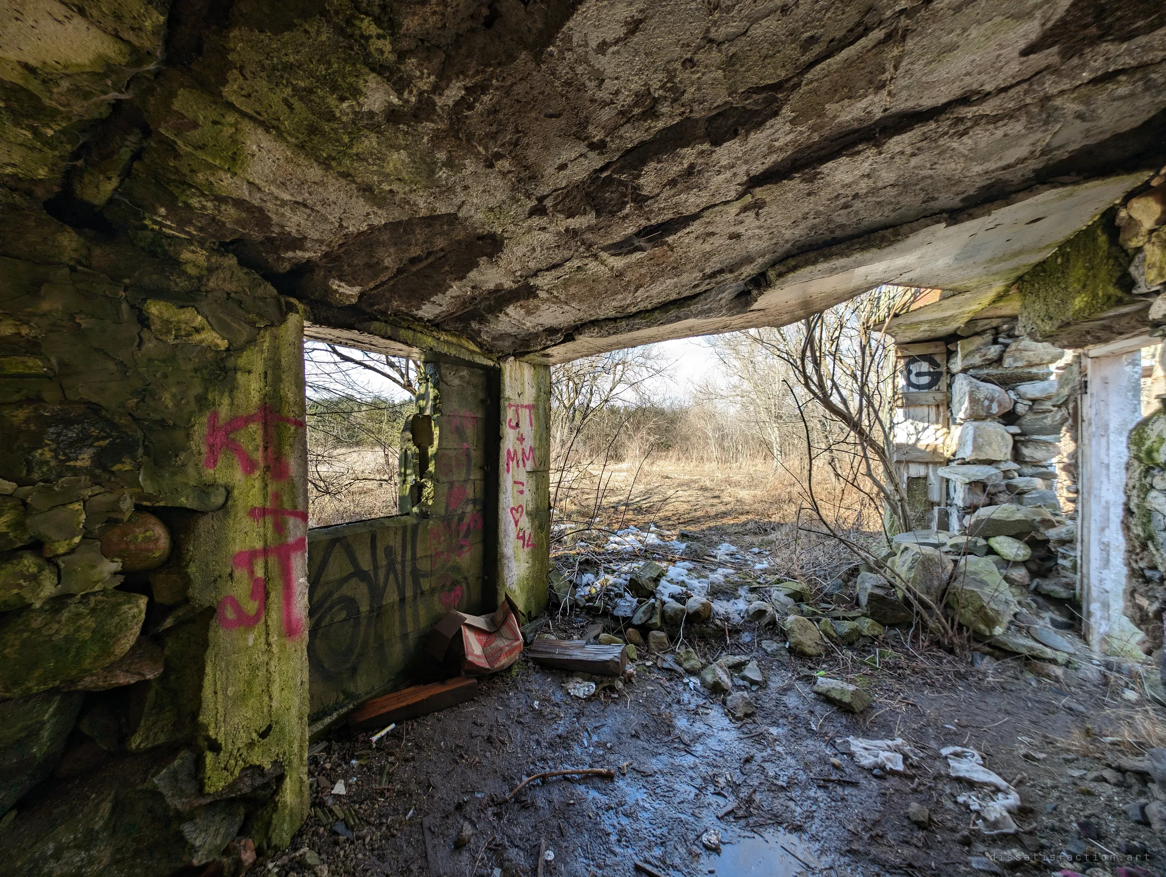 Looking out from under a heavy, moss-stained concrete slab roof, past pillars tagged with initials and hearts, to a field of dry grass and bare trees. A stenciled letter marks the stone wall to the right.