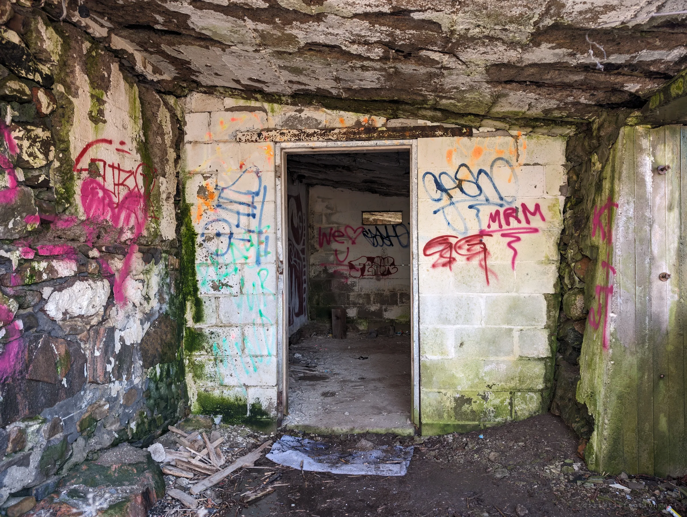 A doorway in a whitewashed block wall under a sagging concrete roof, both sides crowded with graffiti and love notes. Through it, a small bare room with a single slot window.