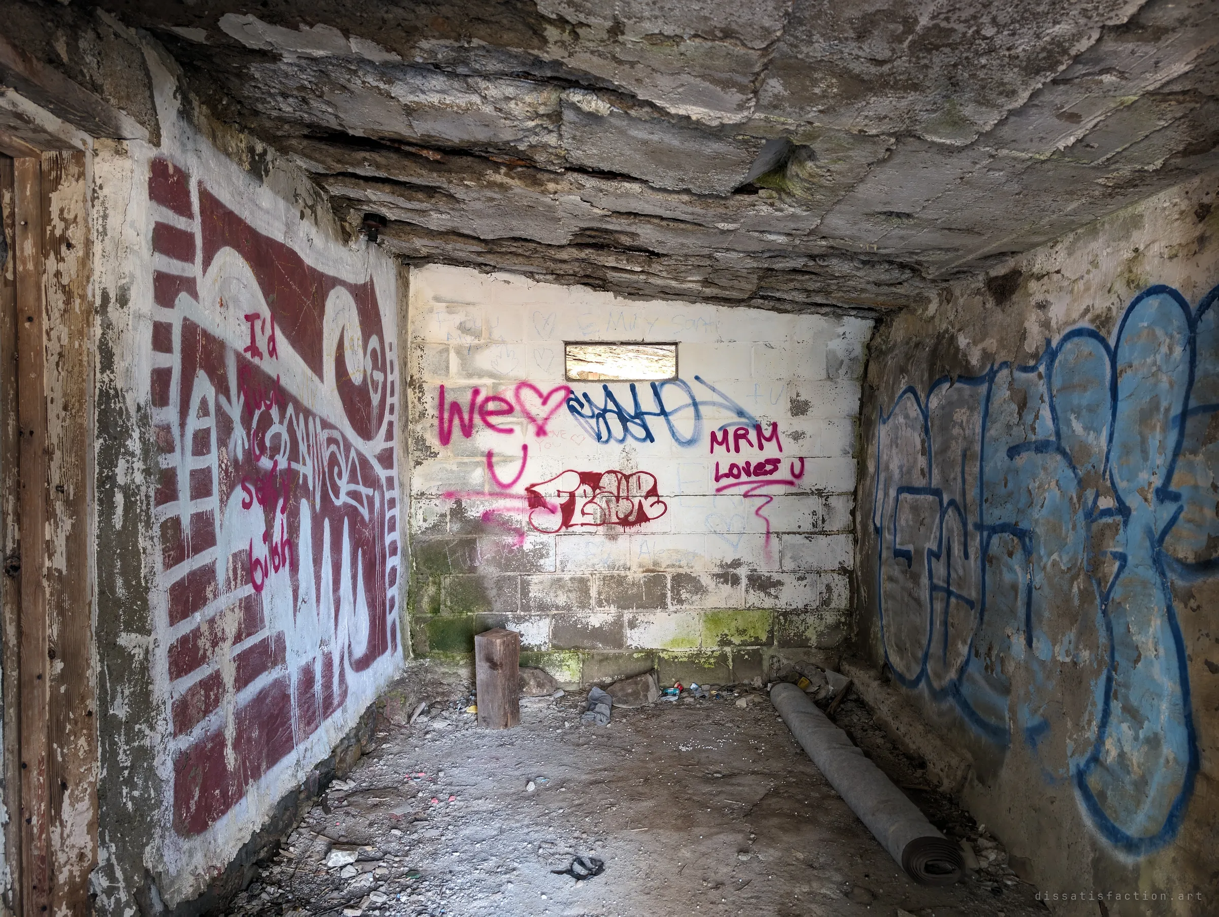 Inside a small concrete cell with a flaking slab ceiling, a slot window high in the back wall and graffiti on every surface, including a large maroon mural and blue letters. A rolled carpet and a wooden block lie on the rubble floor.