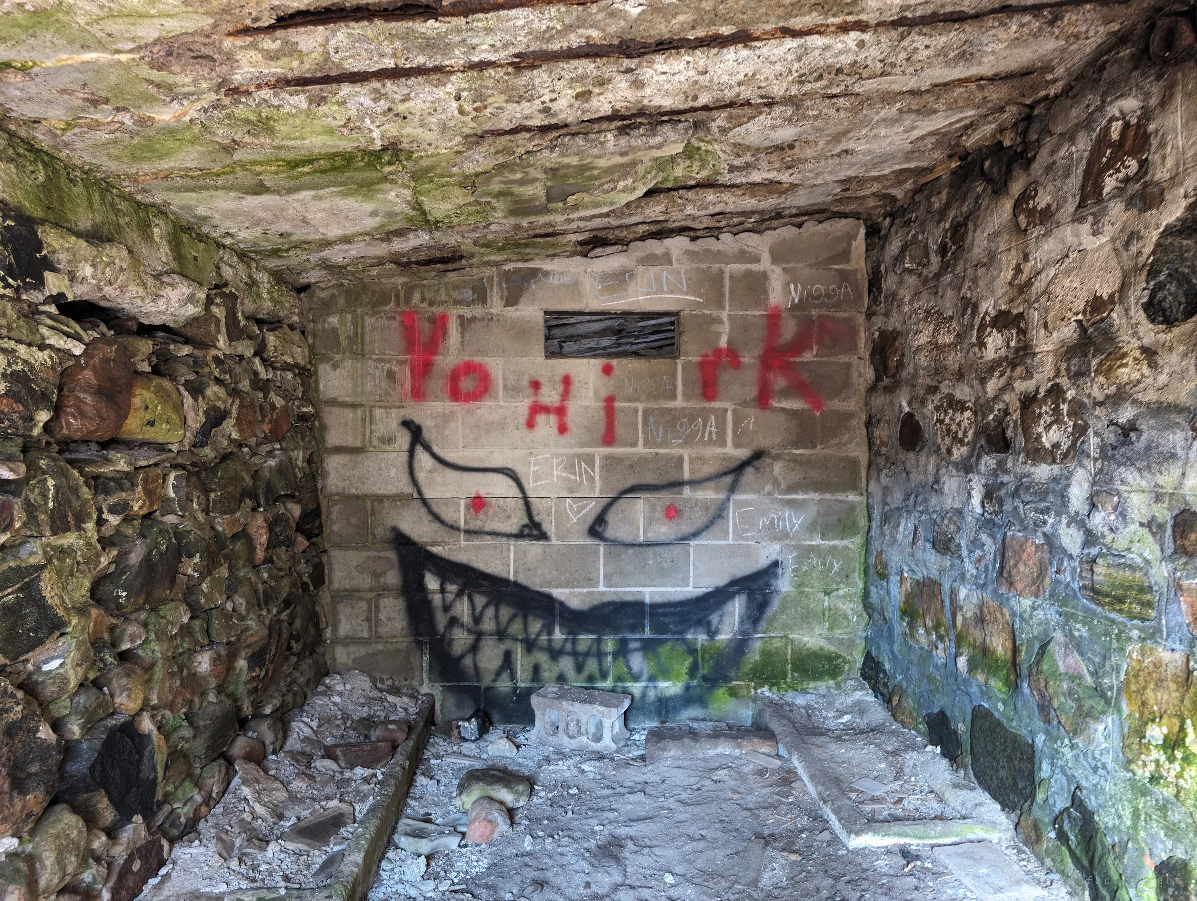 A cramped stone cell with a boarded slot window, its back wall painted with a huge grinning face with red-dot eyes. Scratched names and hearts cover the blocks around it.