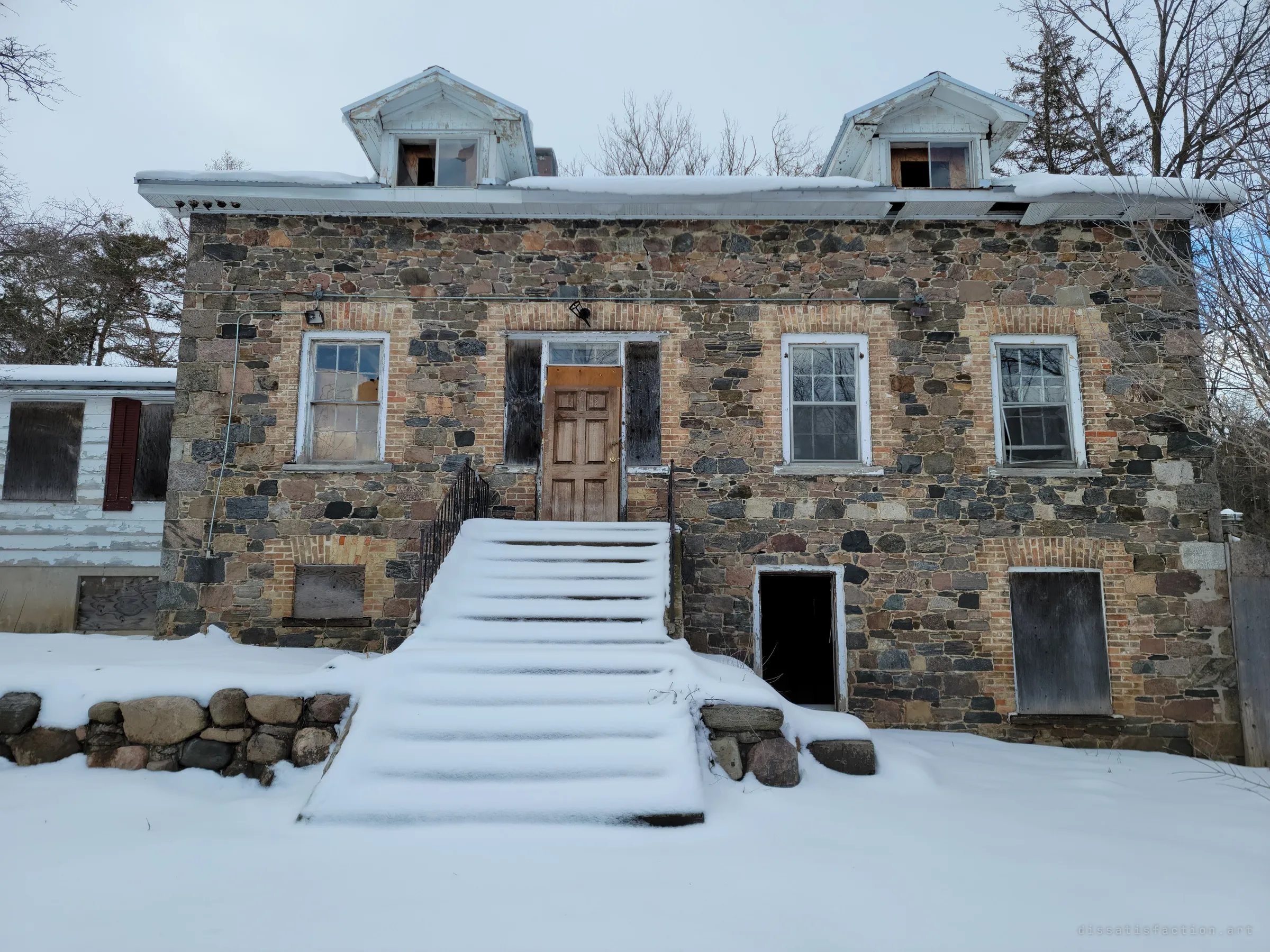 The boarded-up stone farmhouse from the front, its snow-buried steps leading to a patched wooden door under two gabled dormers. It sets the scene: a solid old home left to the winter.