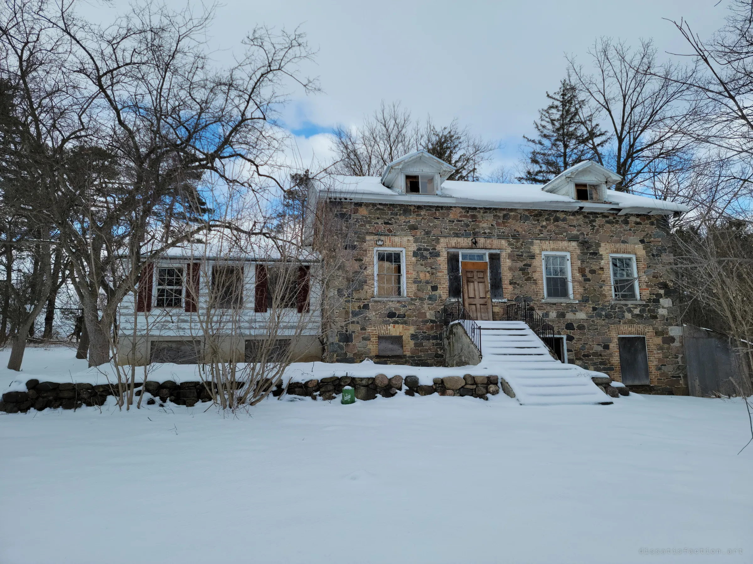 The whole house seen across a snowy yard: a fieldstone main block beside a white clapboard wing with red shutters, framed by bare trees. It shows how the home sits alone in the snow.
