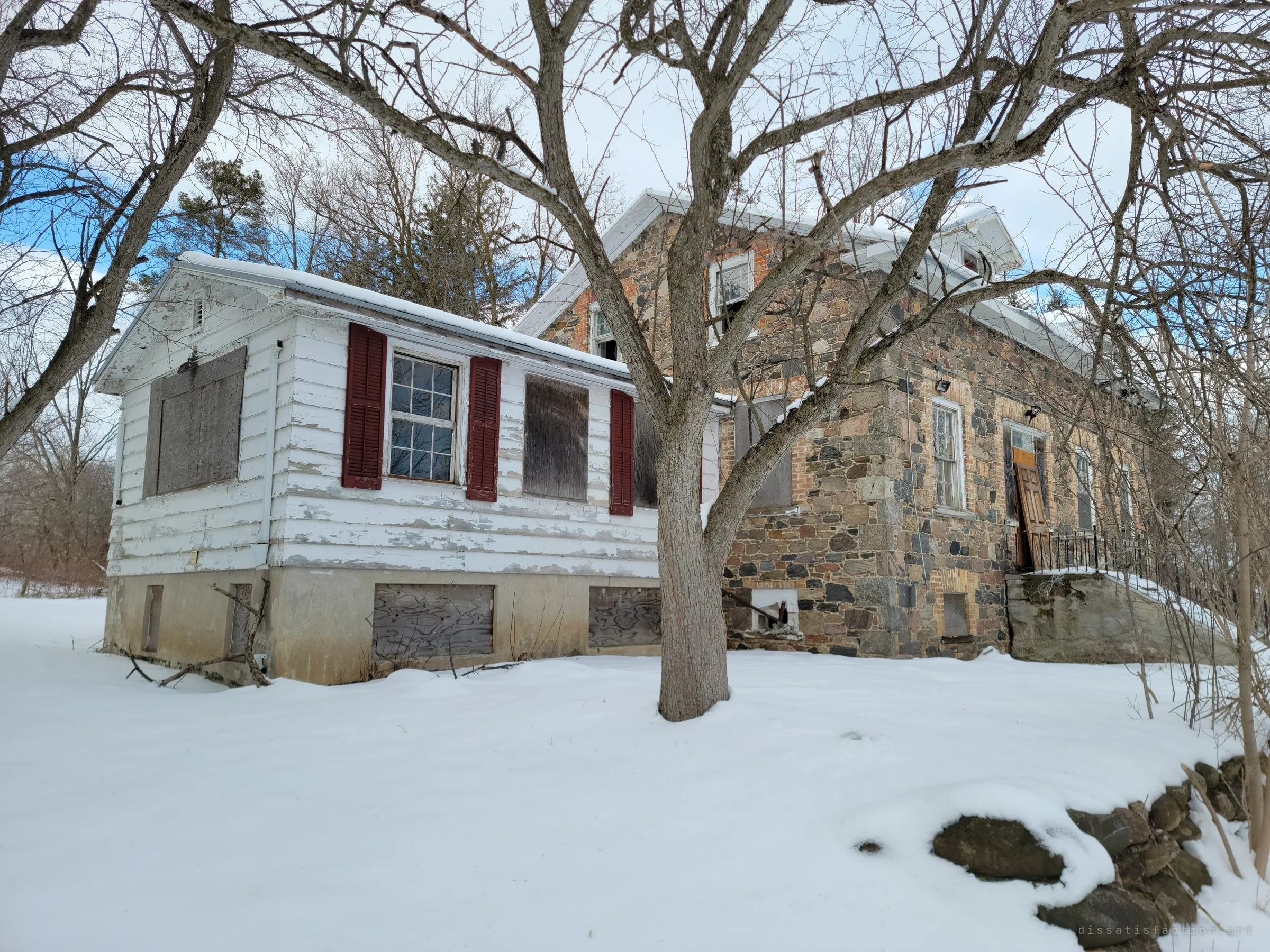 The clapboard wing and stone house from the side, seen through the branches of a bare tree, windows boarded and paint peeling. The low angle makes the empty house loom.