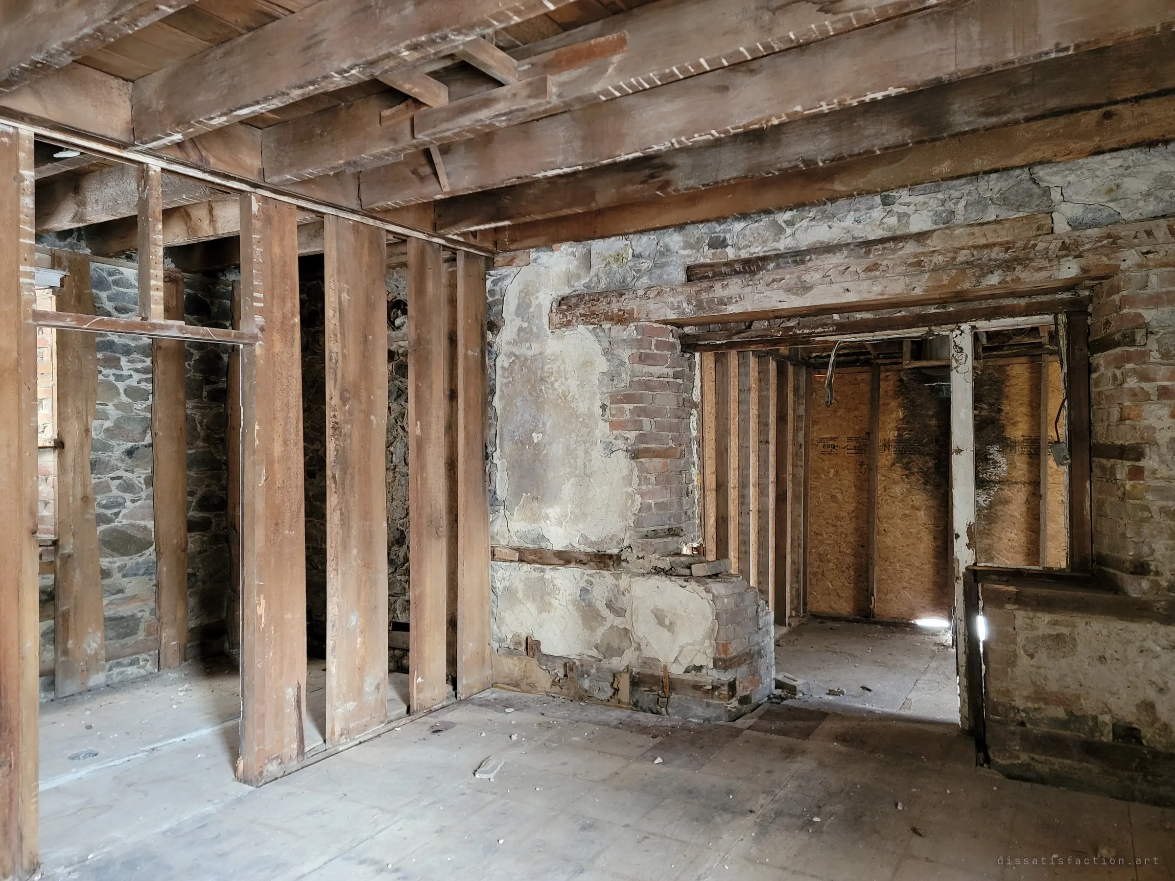 A gutted ground floor with bare stud walls, hand-hewn beams and a crumbling brick chimney against old stone walls. The house has been stripped back to its bones.