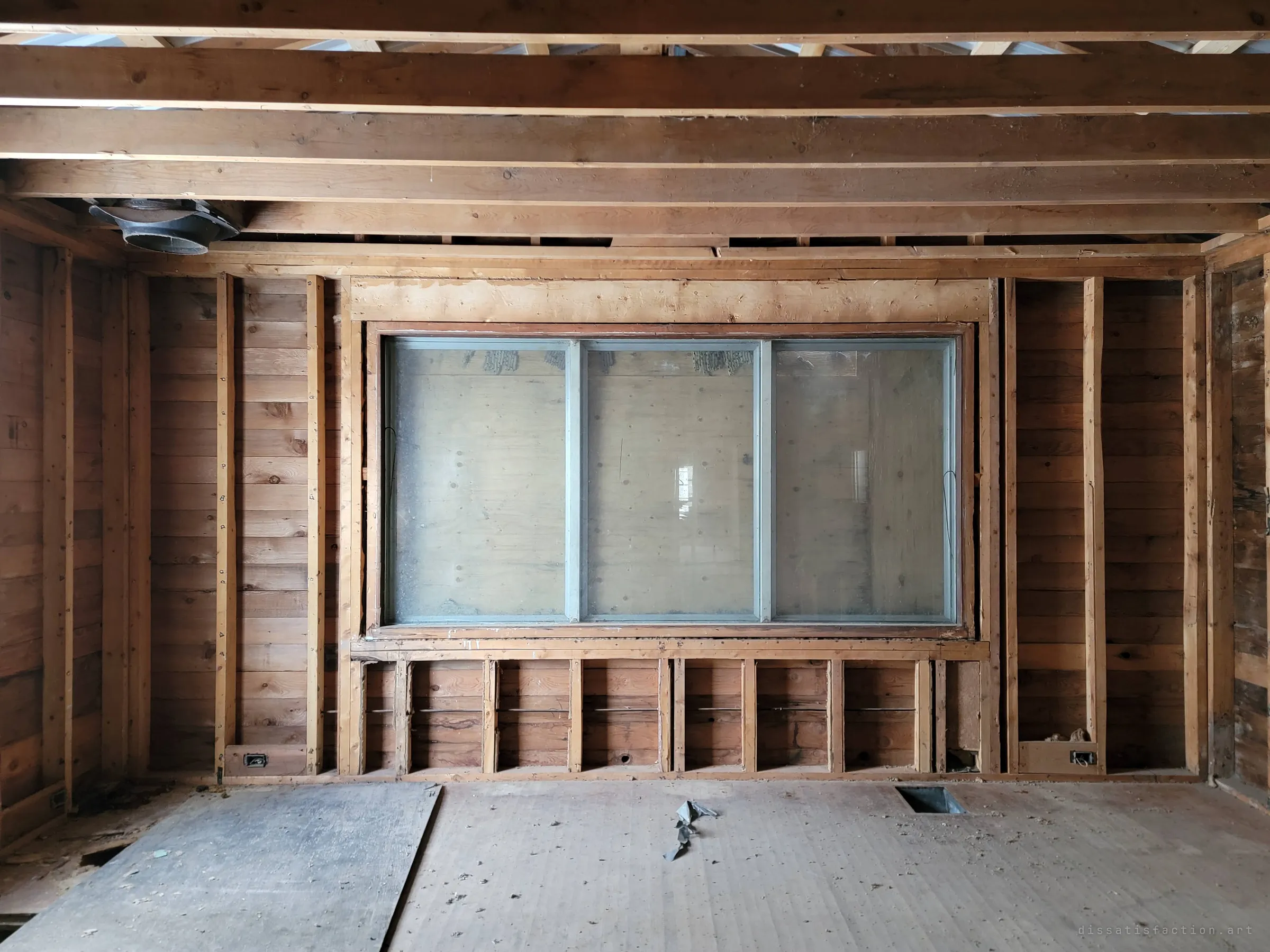 A stripped room of bare plank walls and ceiling joists, centred on a wide three-pane window boarded over from outside. Symmetrical and still, like a stage with no play.