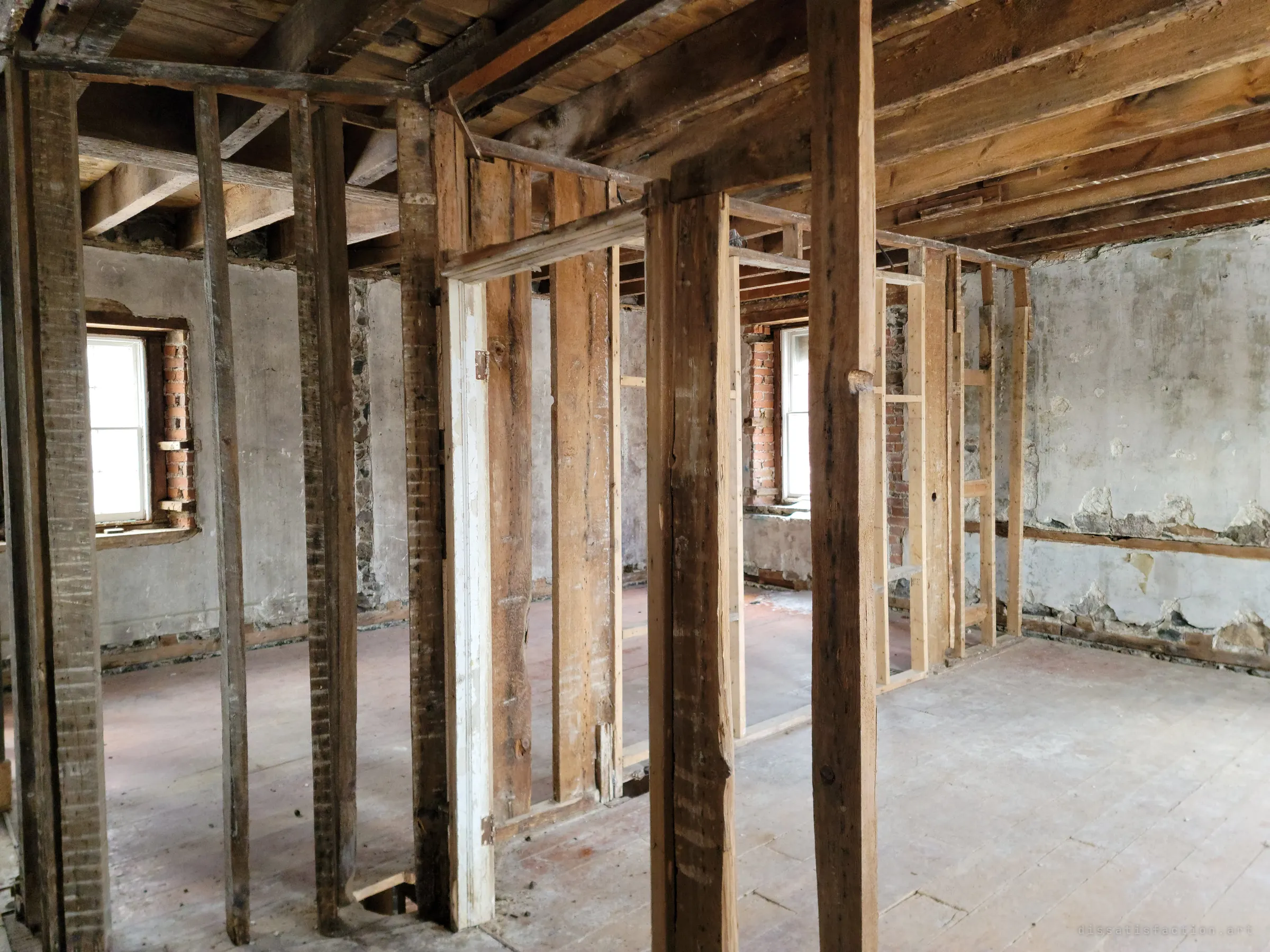 Rough timber posts and stud frames stand in an empty room with plastered stone walls and deep-set windows. Only the skeleton of the old partitions is left.