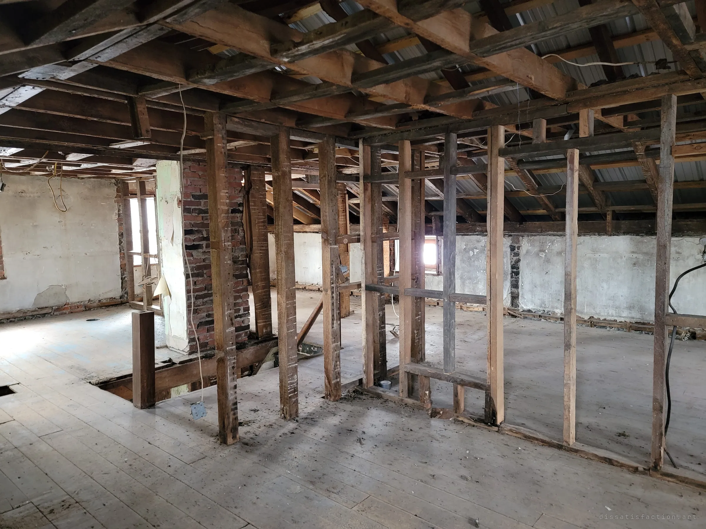 The upper floor opened to the rafters and metal roofing, with bare partition frames, a brick chimney and dangling wires. Light leaks in through a small dormer window.