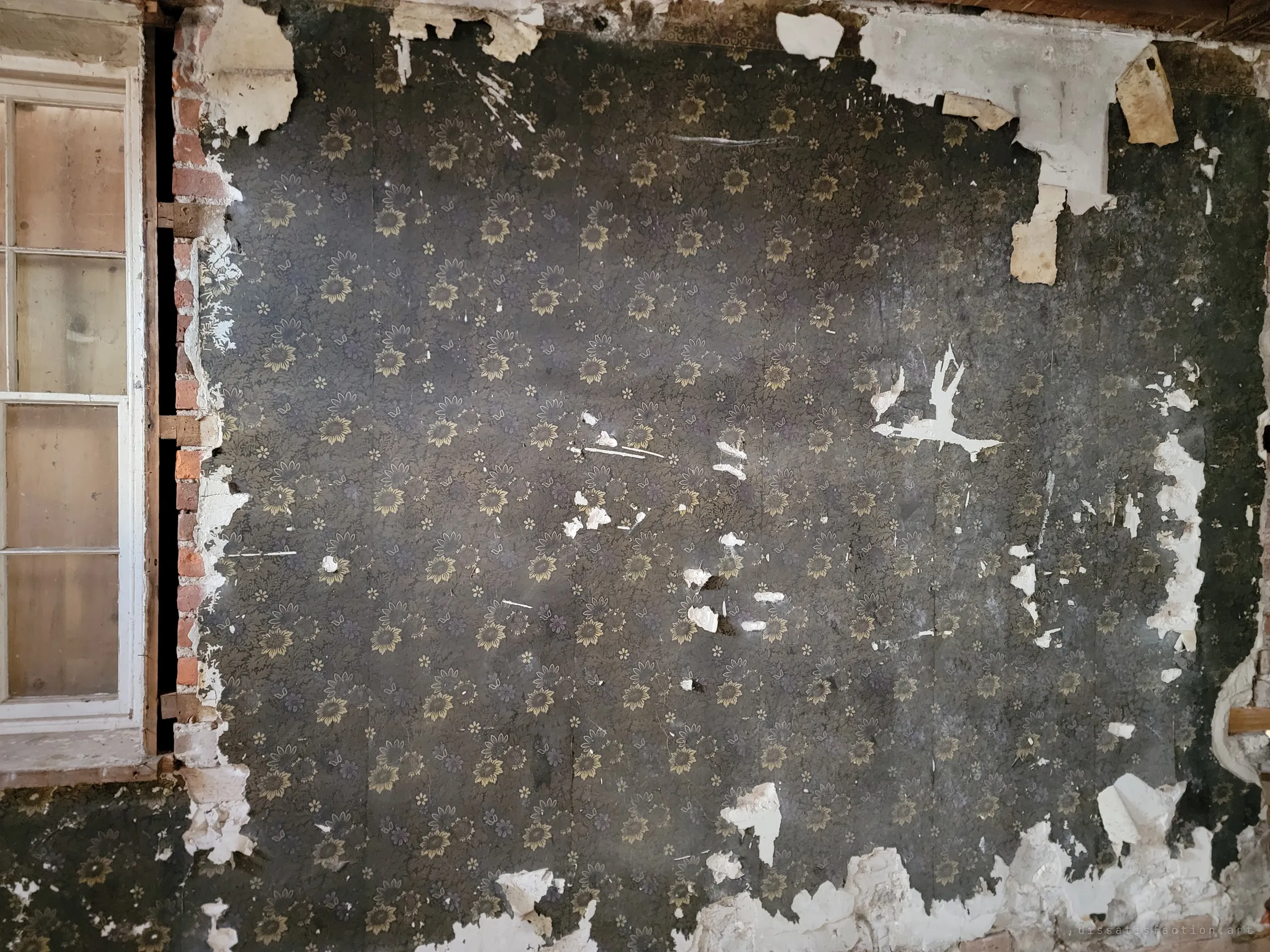 Dark floral wallpaper peeling away from plaster and brick beside a tall window. The old decoration still clings on in patches.