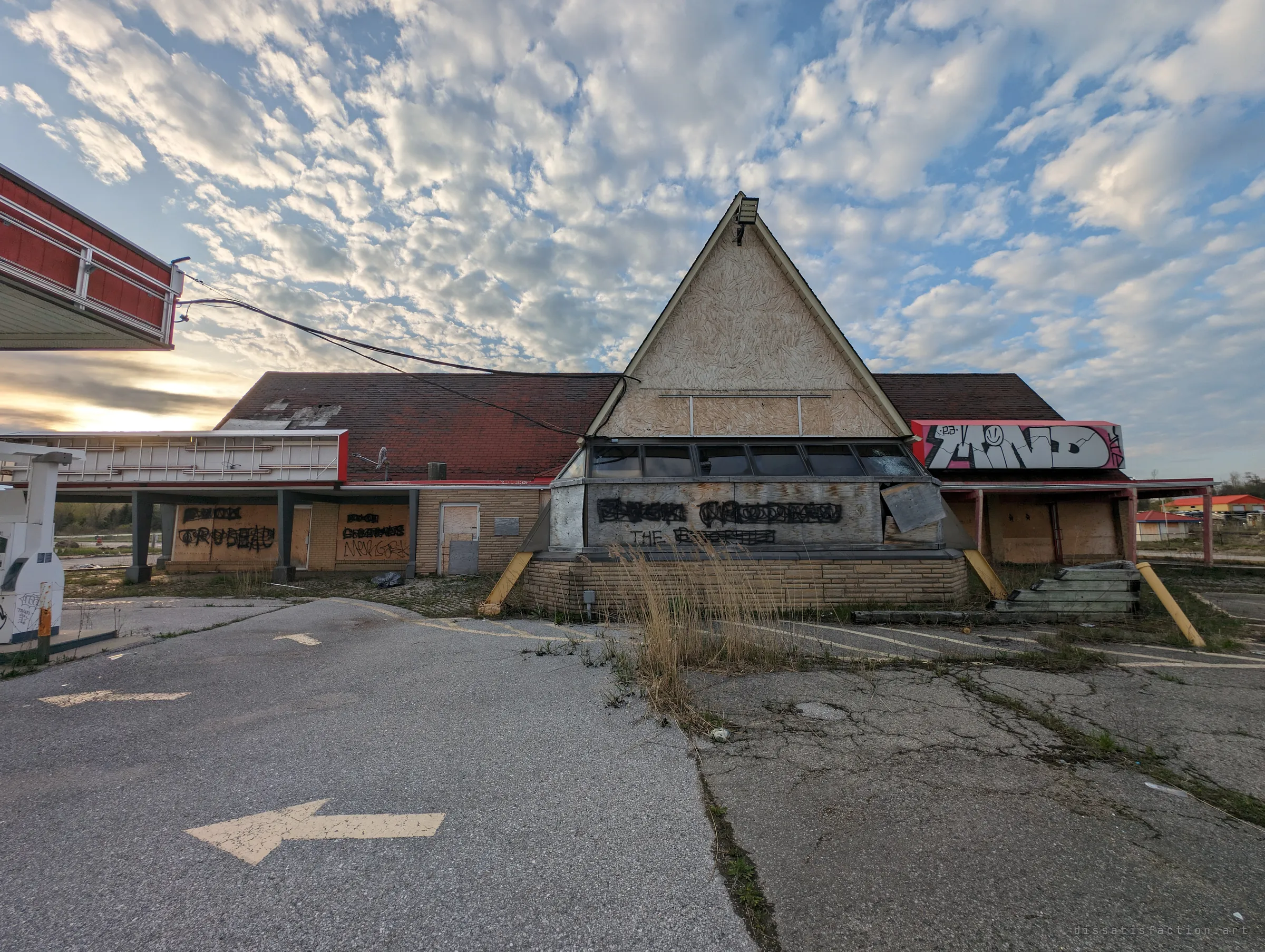 A boarded-up A-frame roadside restaurant under a mackerel sky, graffiti across its plywood and weeds pushing through the cracked lot. The whole place has been left to the weather.