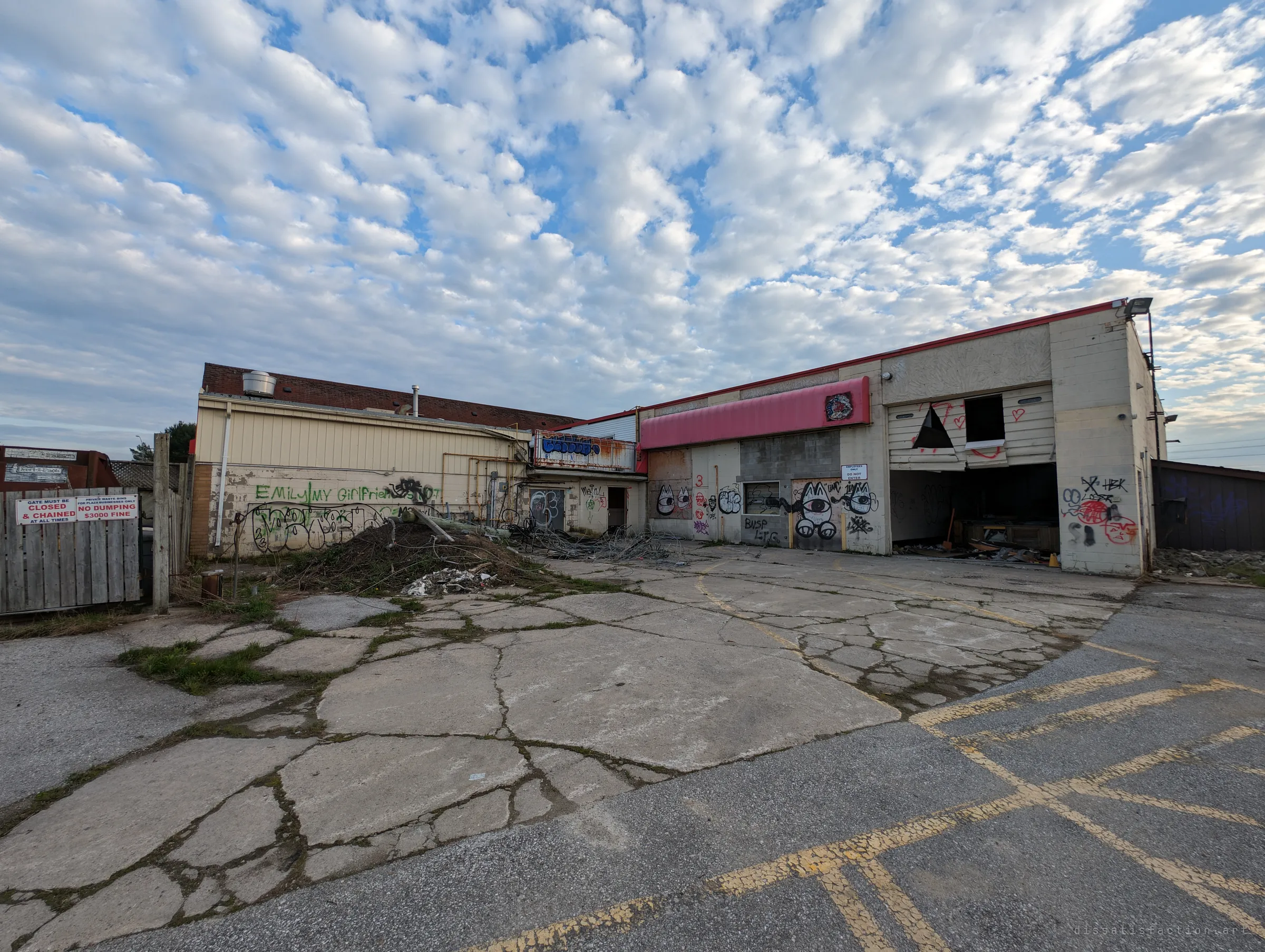 The back of the plaza: a gutted service bay with its door smashed open, walls layered in graffiti and a cracked concrete yard going green.