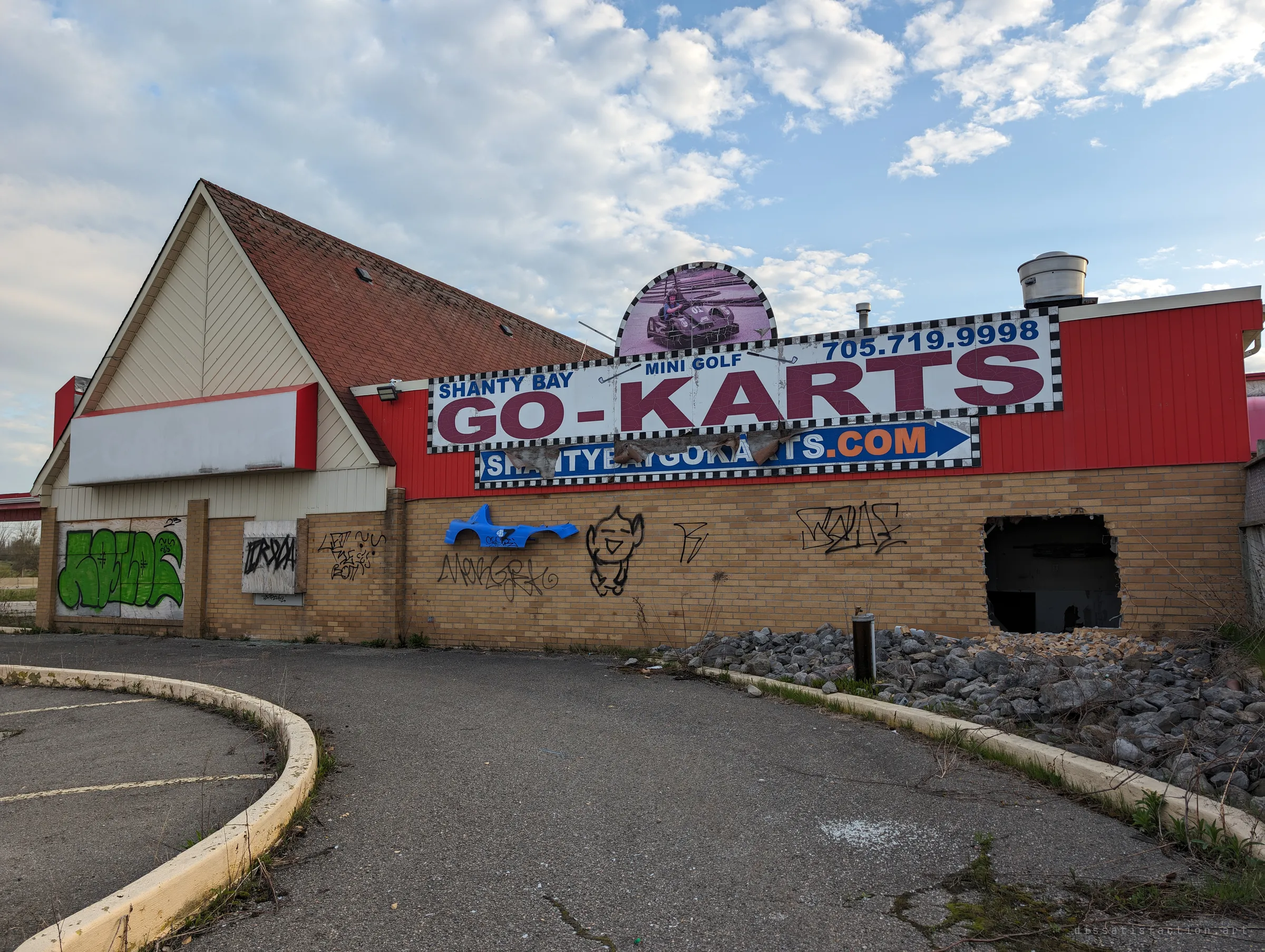 The faded go-kart and mini-golf sign still hangs over a graffiti-tagged brick wall, a broken kart body pinned beside it and a hole punched through the bricks.