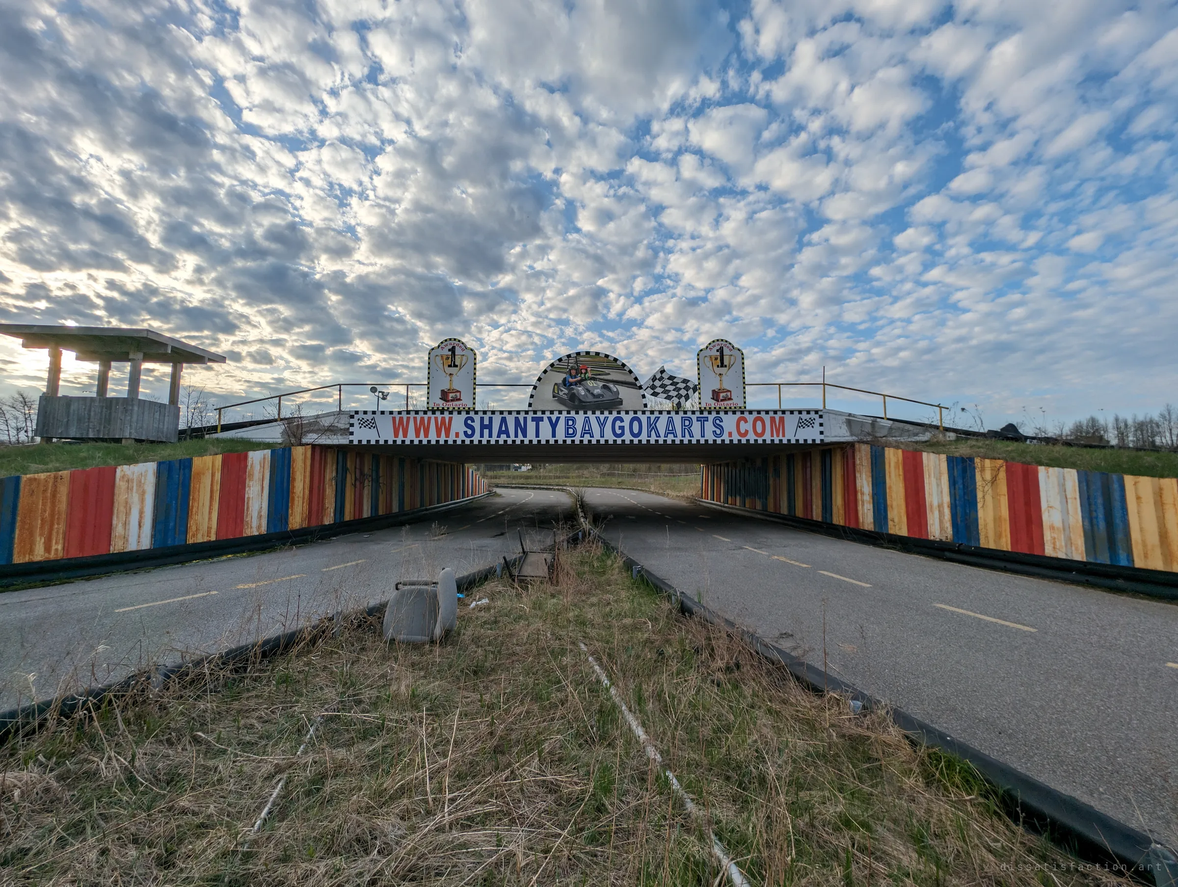 The go-kart track splits beneath a striped overpass still dressed in racing banners, a lost seat sitting in the overgrown median.