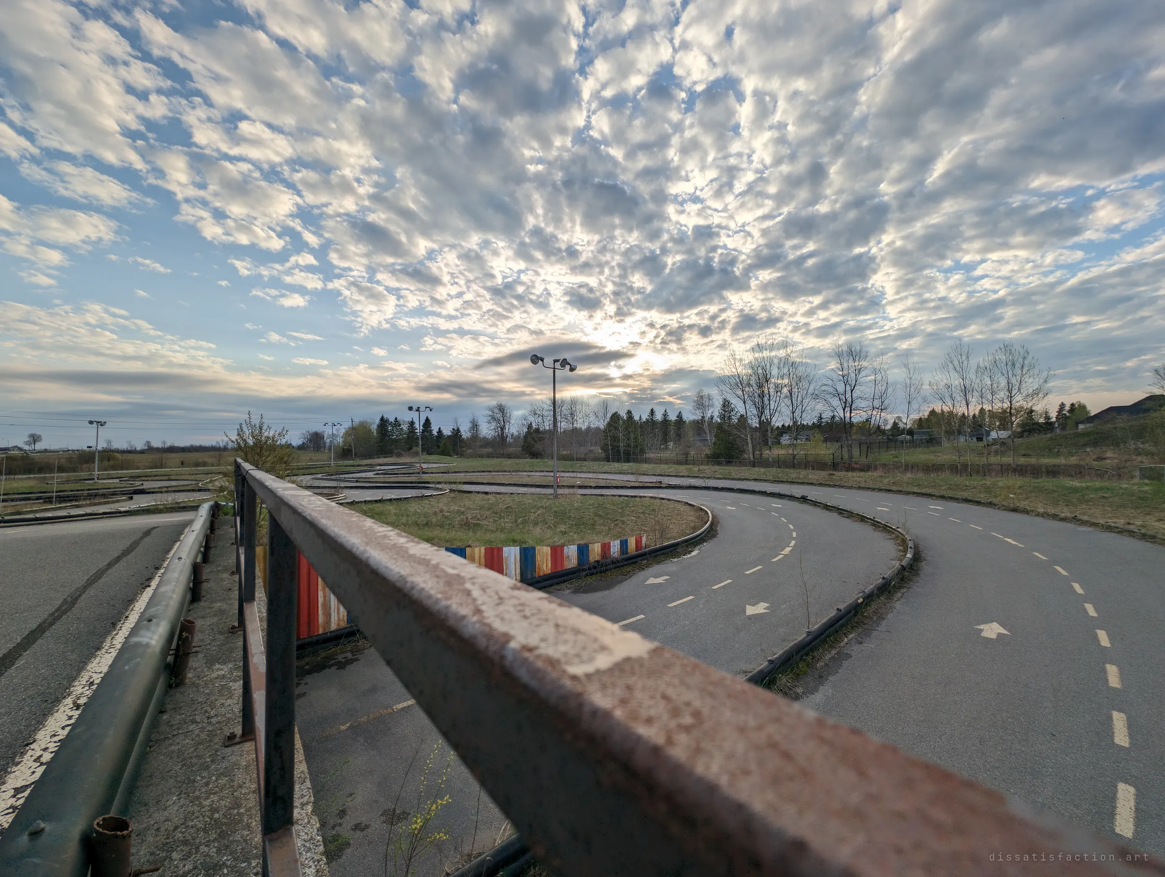 From the rusted bridge rail, the empty kart track curls away through dry grass toward a low sun, arrows still pointing the way no one drives.