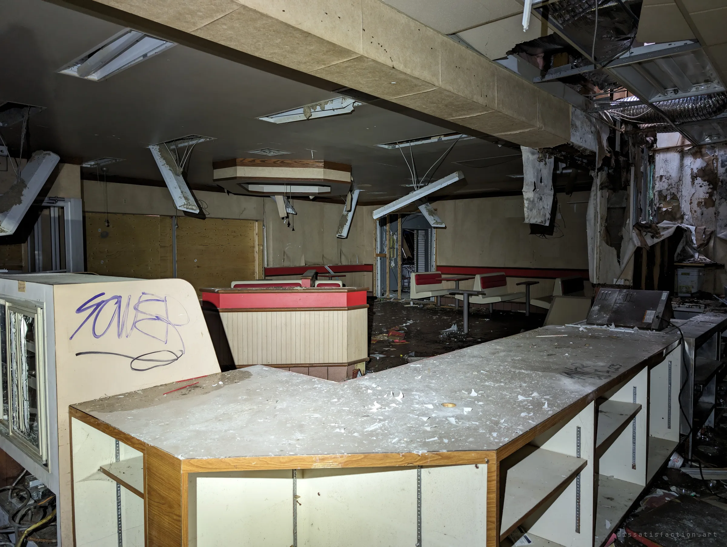 A dust-covered service counter in a gutted fast-food dining room, ceiling panels and light fittings hanging loose over red-trimmed booths. The retro booths and fallen ceiling show how long the place has sat empty.