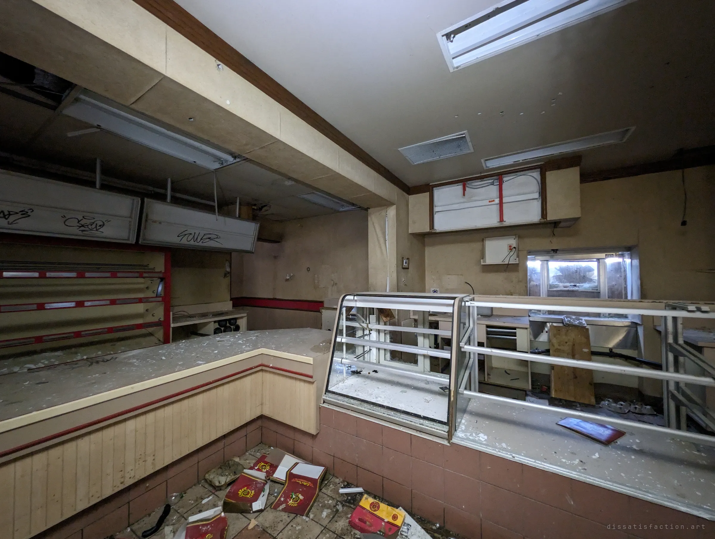 Behind the counter of an abandoned coffee shop: an emptied pastry case, bare display racks and branded boxes scattered across the tiles, with a clouded pickup window at the back.