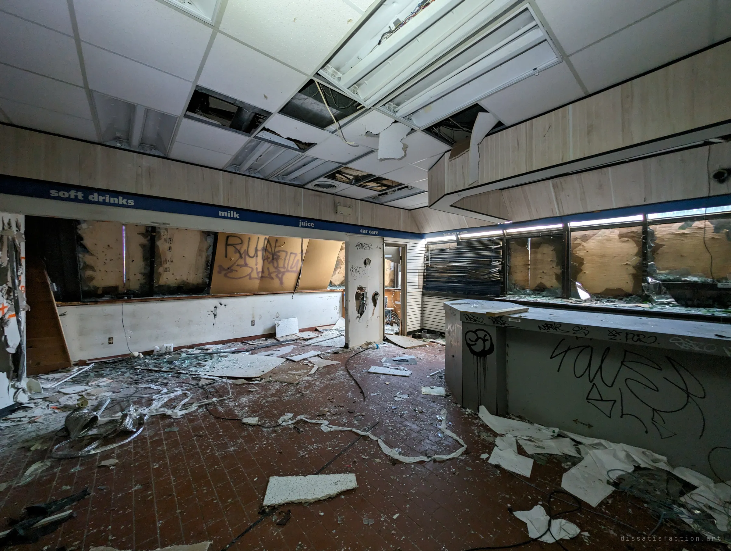 A wrecked convenience store with its old aisle banner still running along the wall above boarded windows, the floor buried in glass, paper and ceiling tiles.