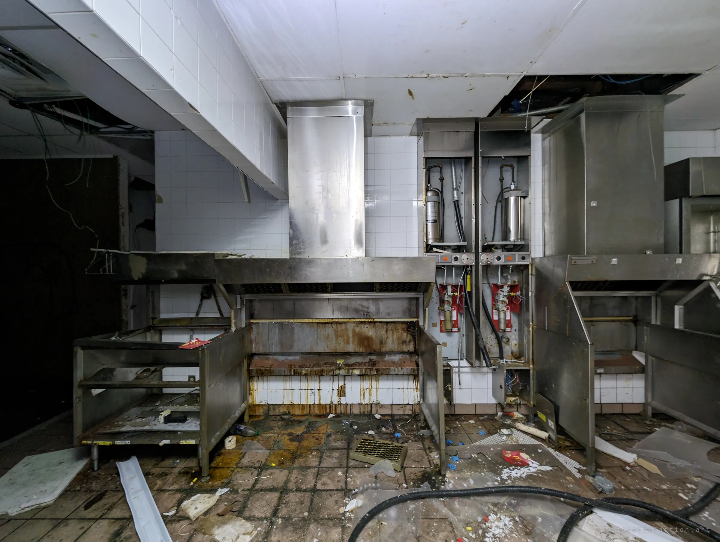 A stripped fast-food kitchen line: steel hoods and fire-suppression tanks remain, but the fryers and grills are gone, leaving rust-streaked tile where they stood.