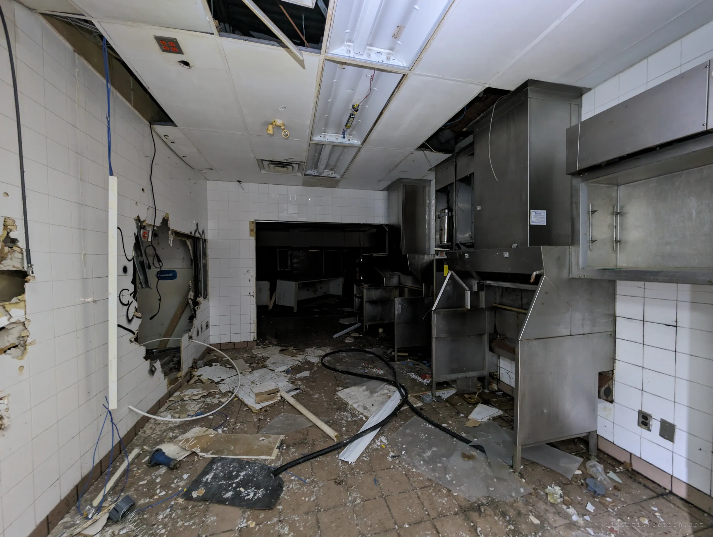 A tiled back kitchen with walls punched open for scrap, hoses and debris across the floor and a dark serving area beyond the doorway.