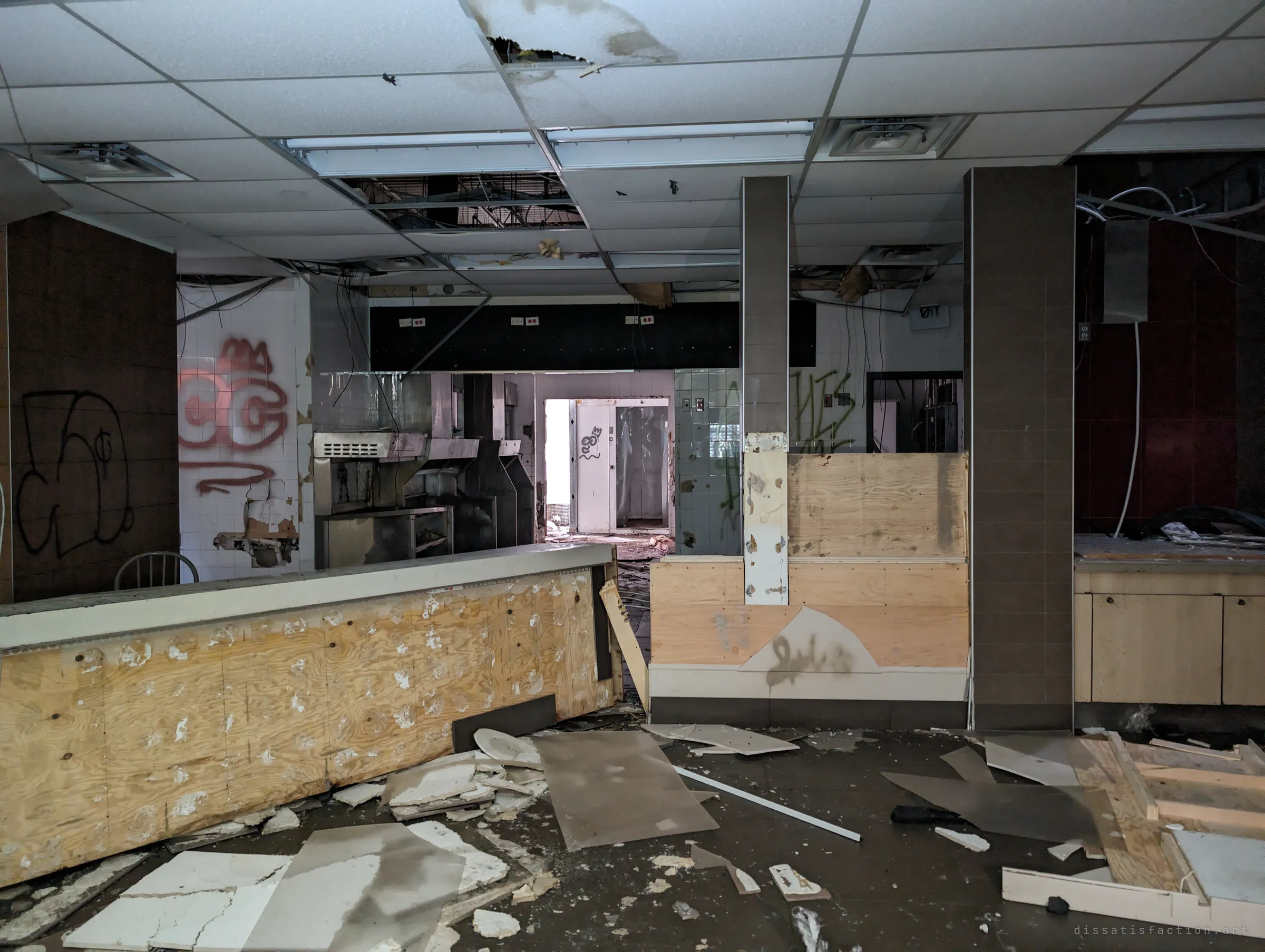A plywood-patched front counter in a graffiti-covered food court, looking through the gutted kitchen toward a bright back door.