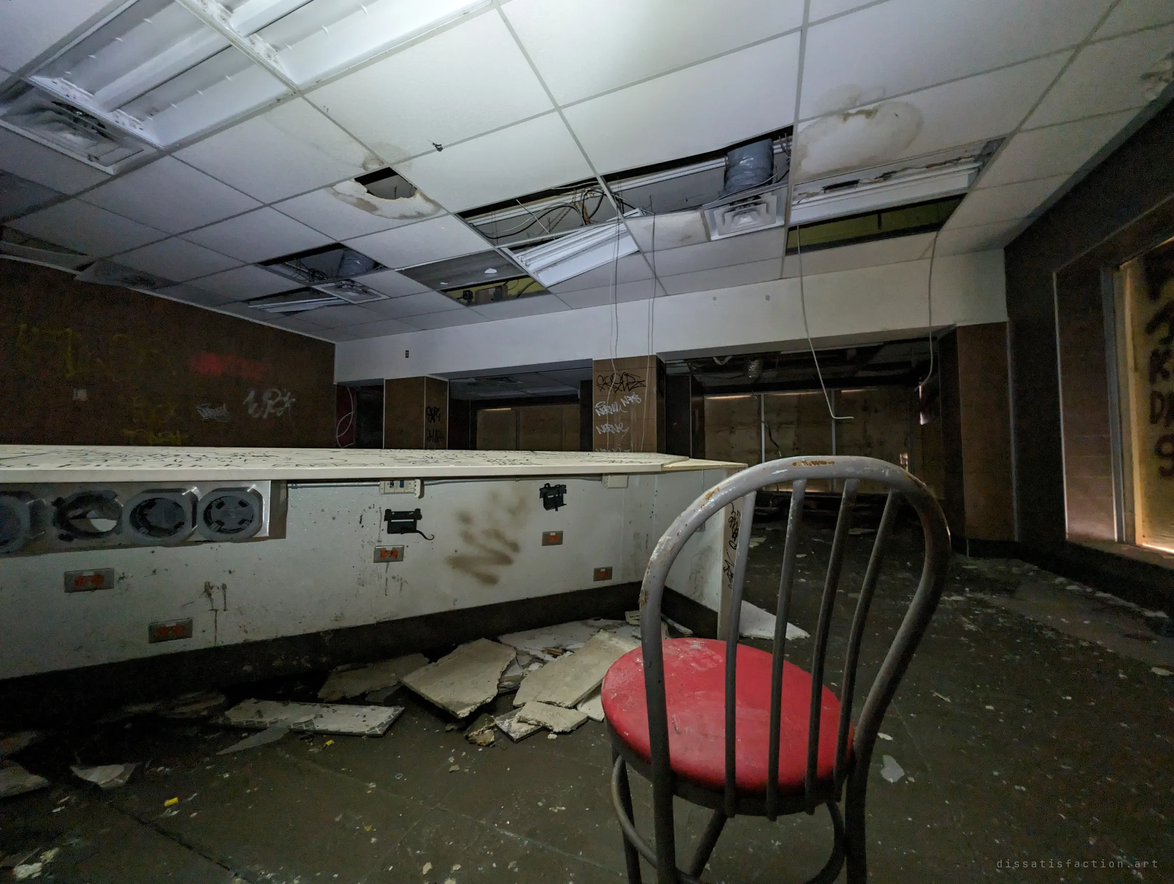 A lone red-seated chair beside a long counter of empty power outlets, under a sagging, water-stained ceiling in the abandoned dining hall.