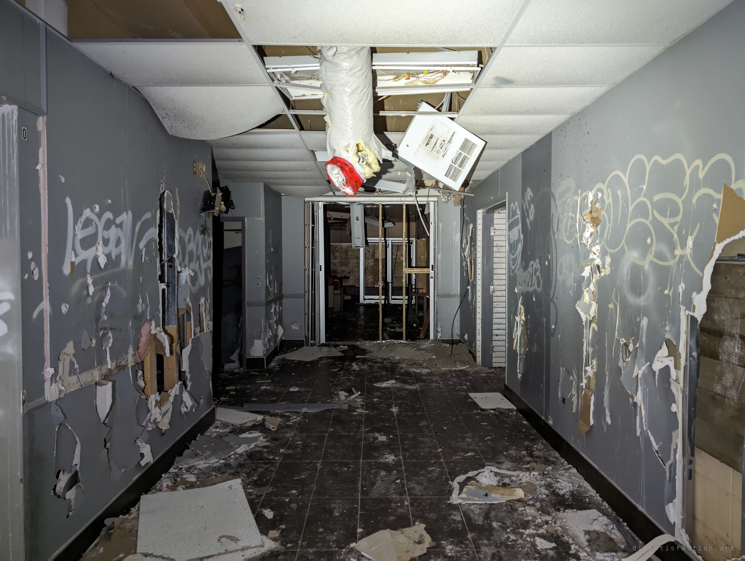 A grey corridor with peeling walls and heavy tags, a torn duct dangling from the ceiling, leading to a wrecked room behind glass doors.