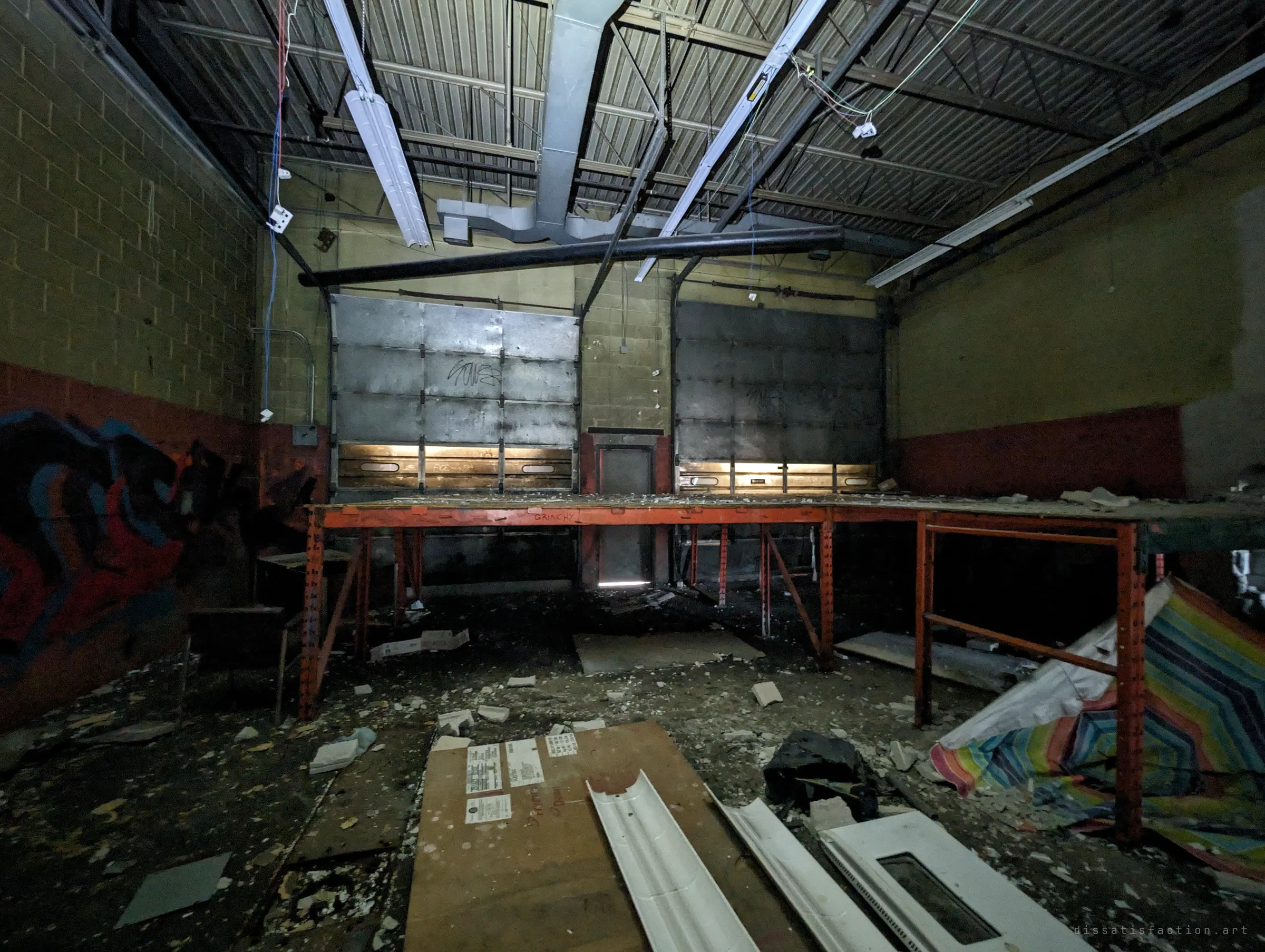 A dark service bay with a rusted orange mezzanine, two roll-up doors glowing at their seals and a torn beach umbrella on the littered floor.