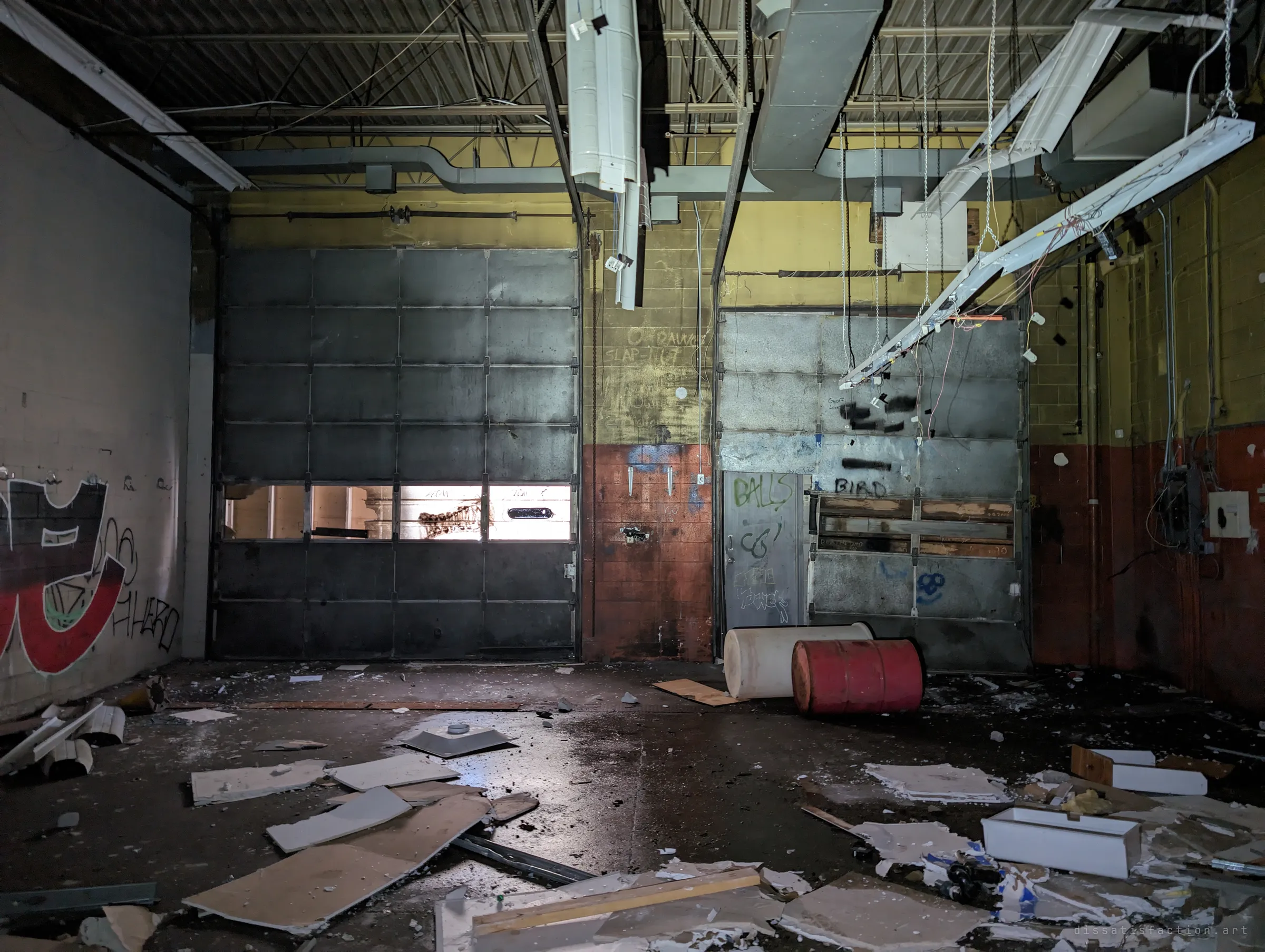 Two tagged roll-up doors in a concrete-block garage bay, daylight leaking through one door's windows onto a floor of fallen ceiling tiles and old drums.