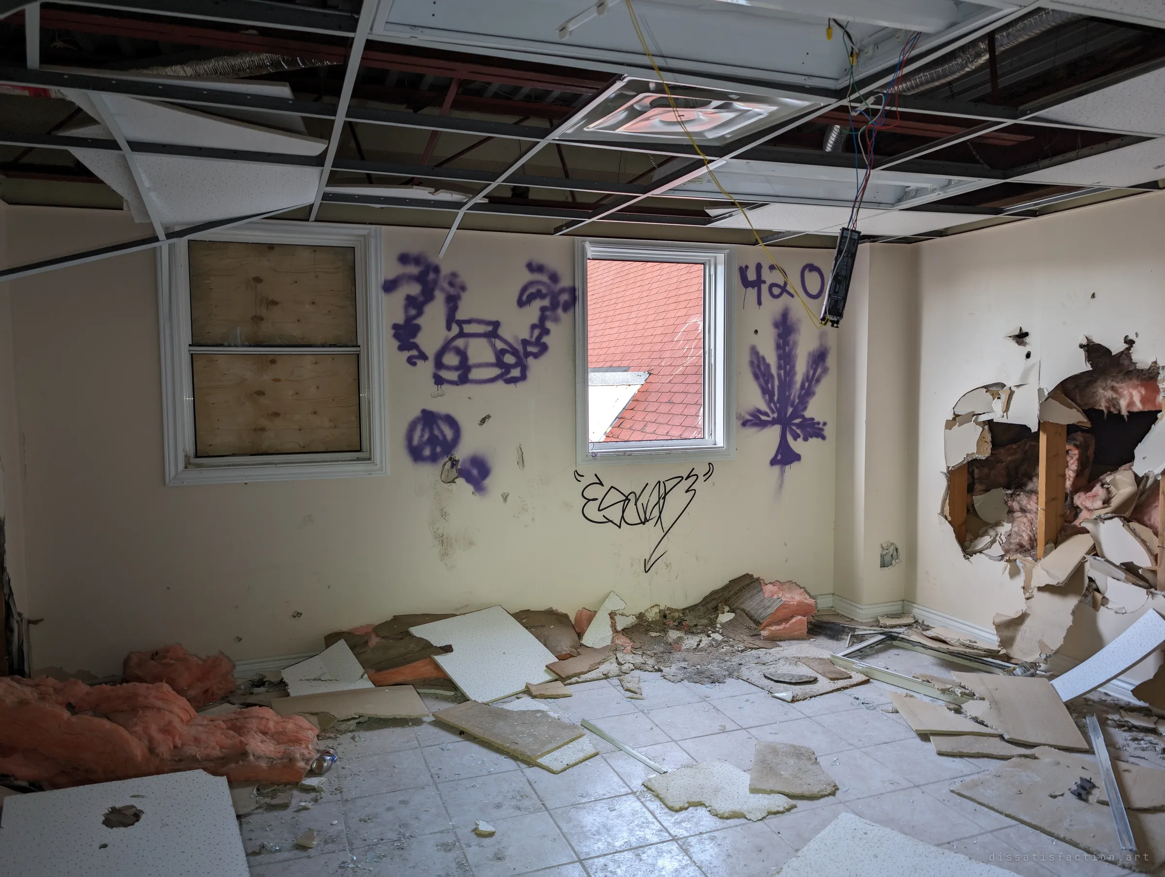 An upstairs room with a collapsed drop ceiling, a boarded window beside an open one showing a red shingled roof, and walls smashed open to the insulation.