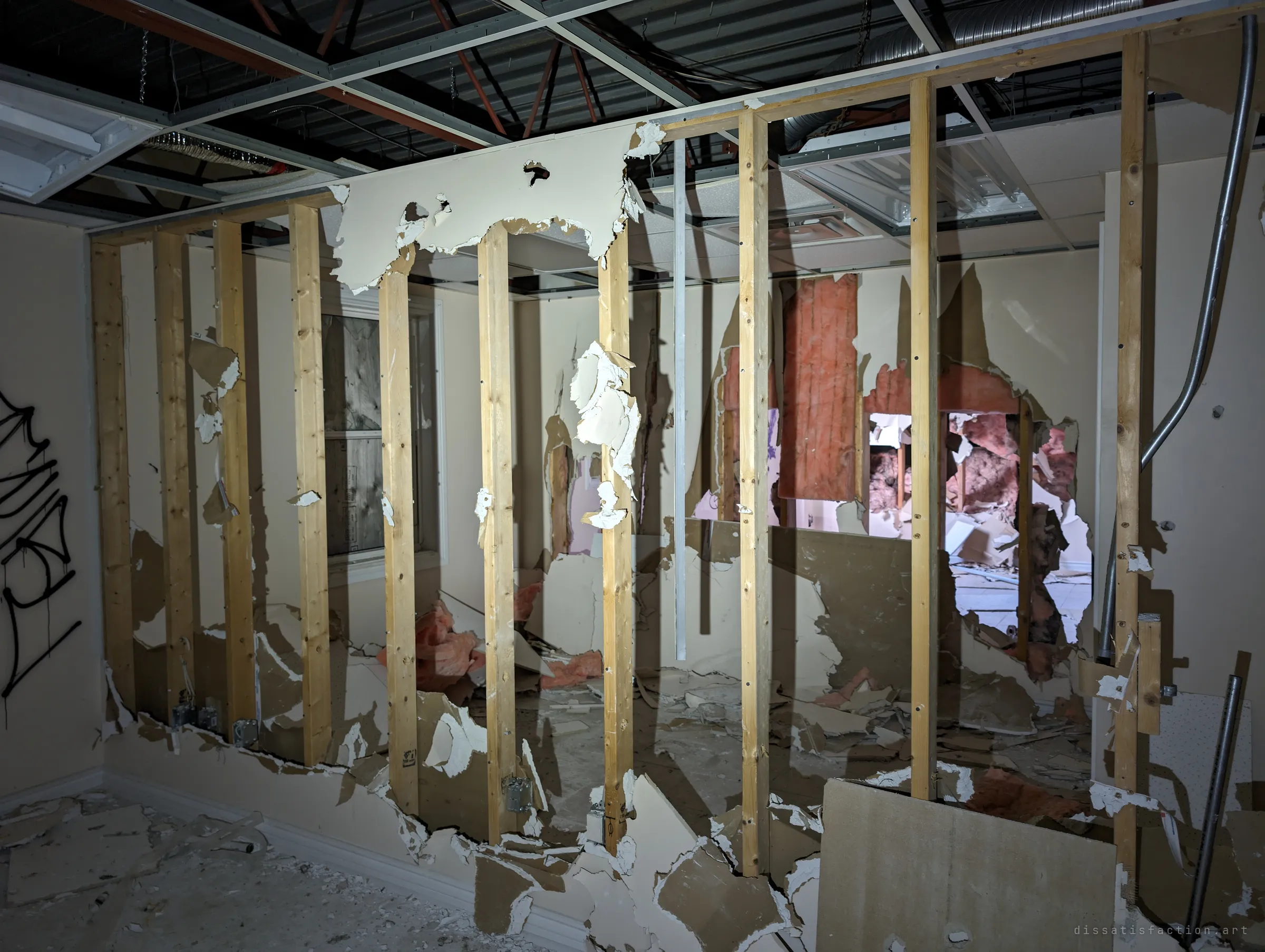 A partition wall stripped to its wooden studs, torn drywall hanging off it and insulation spilling from holes in the rooms beyond. The building is being gutted from the inside out.