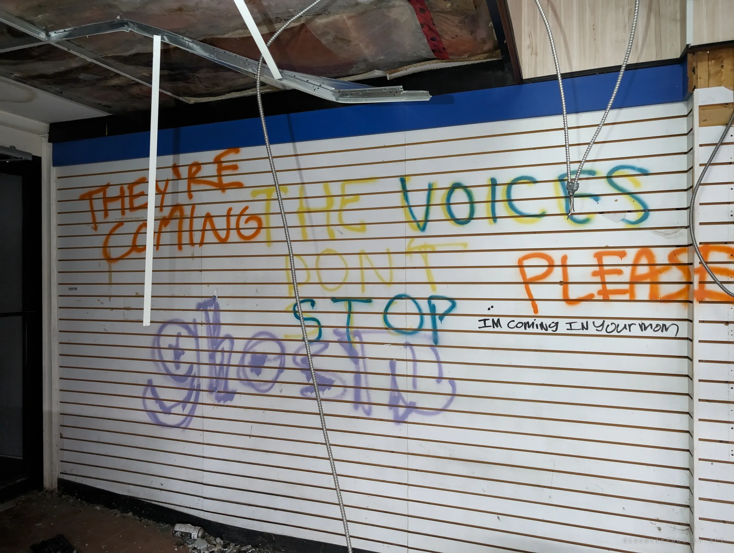A white slatwall display wall under a collapsed ceiling grid, covered in spray-painted pleas like "the voices don't stop" and "please". Loose armoured cables dangle through the frame.