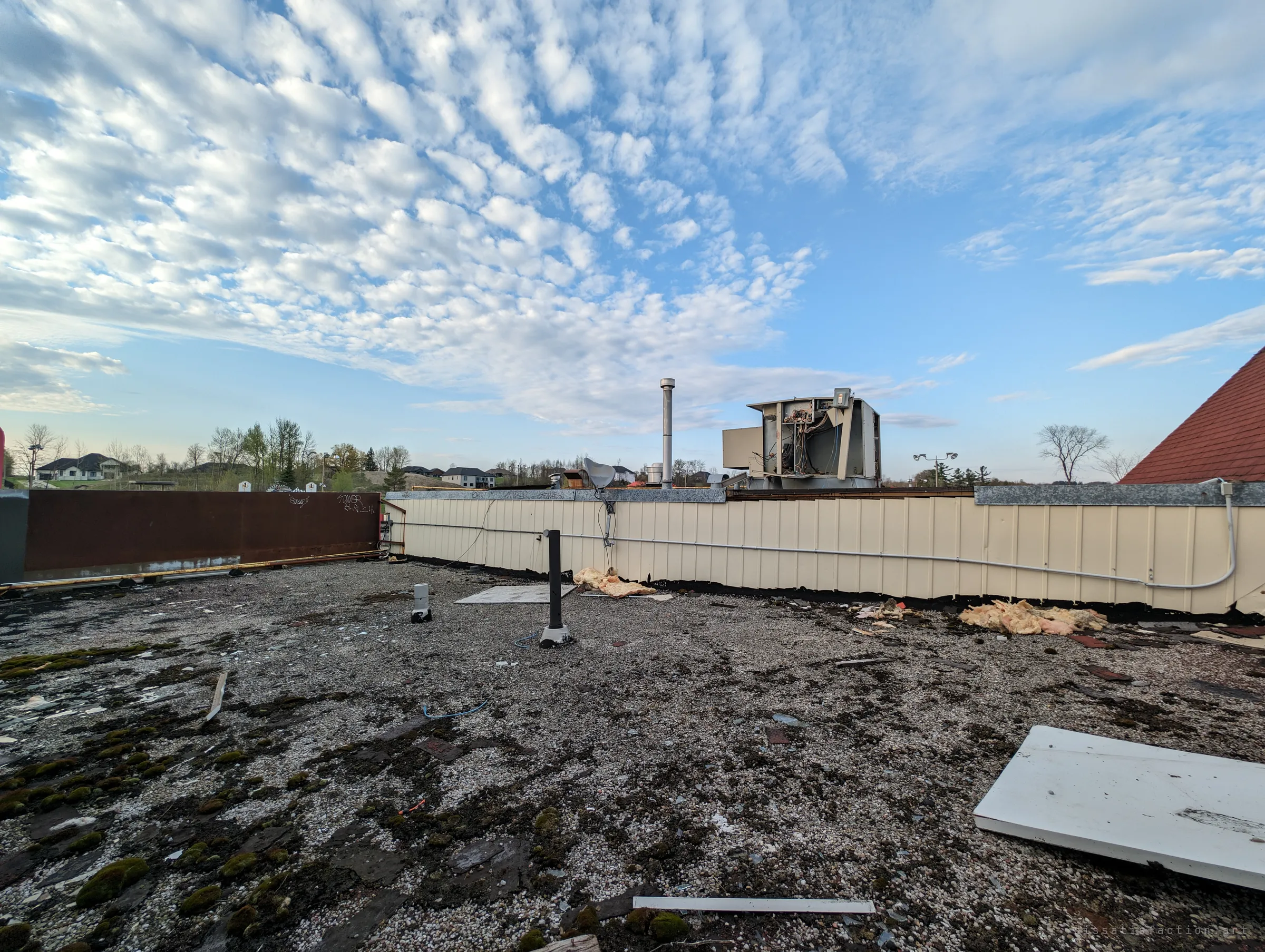 The gravel roof, patched with moss and scattered insulation, under a wide sky of rippled cloud. A gutted rooftop unit sits open to the weather.