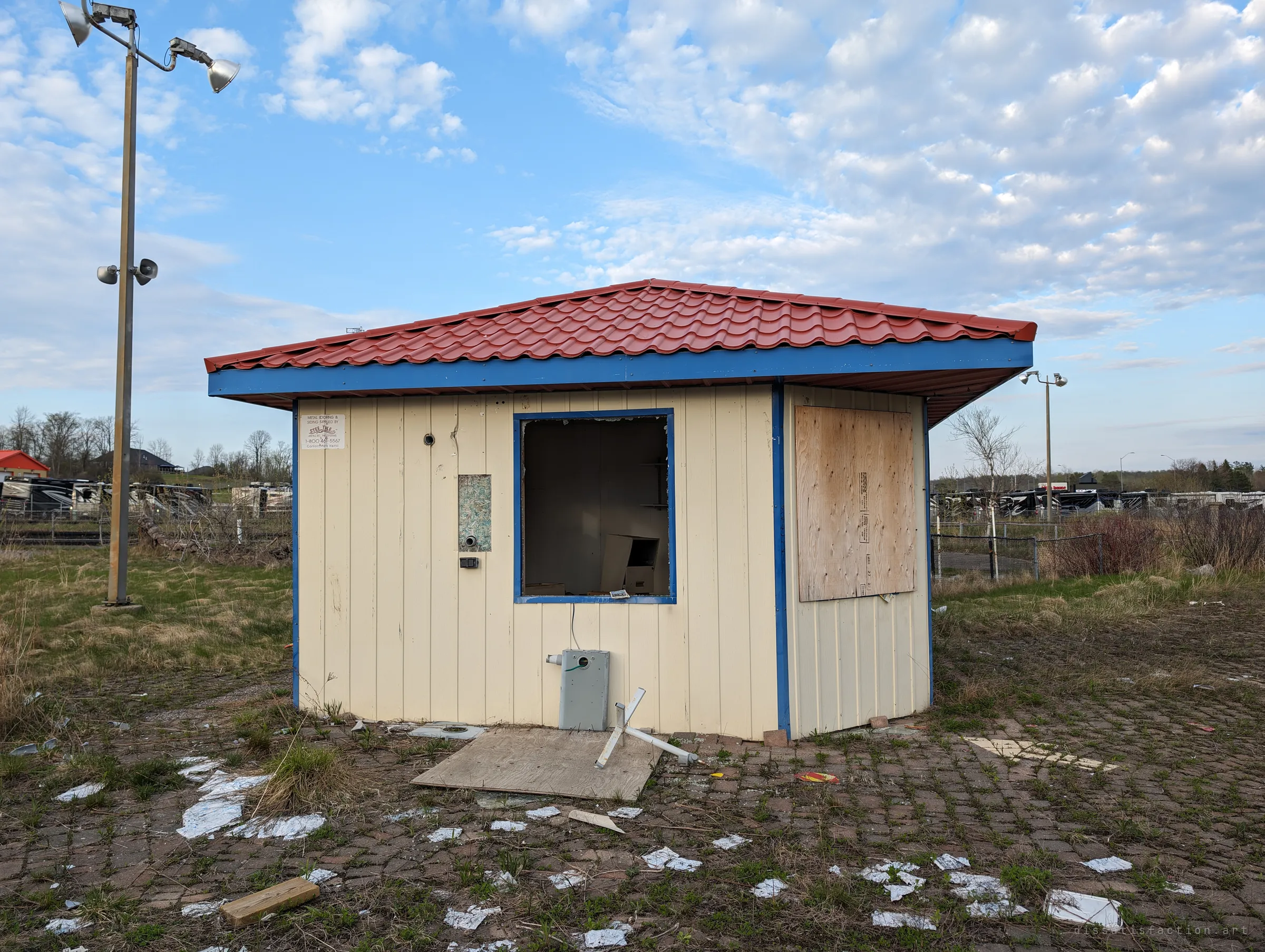 A small ticket booth with a red tile roof and blue trim, its service window smashed open and one side boarded up. Broken tiles and paper litter the weedy paving around it.
