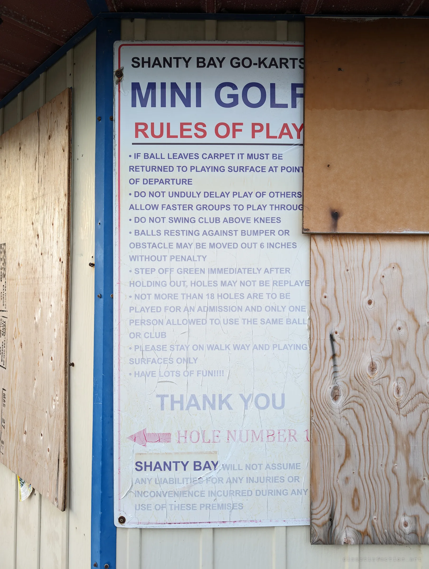 A faded mini golf rules sign between boarded-up windows, its print peeling and cracked. It ends with a cheerful "have lots of fun!!!!" and a warning about liability.