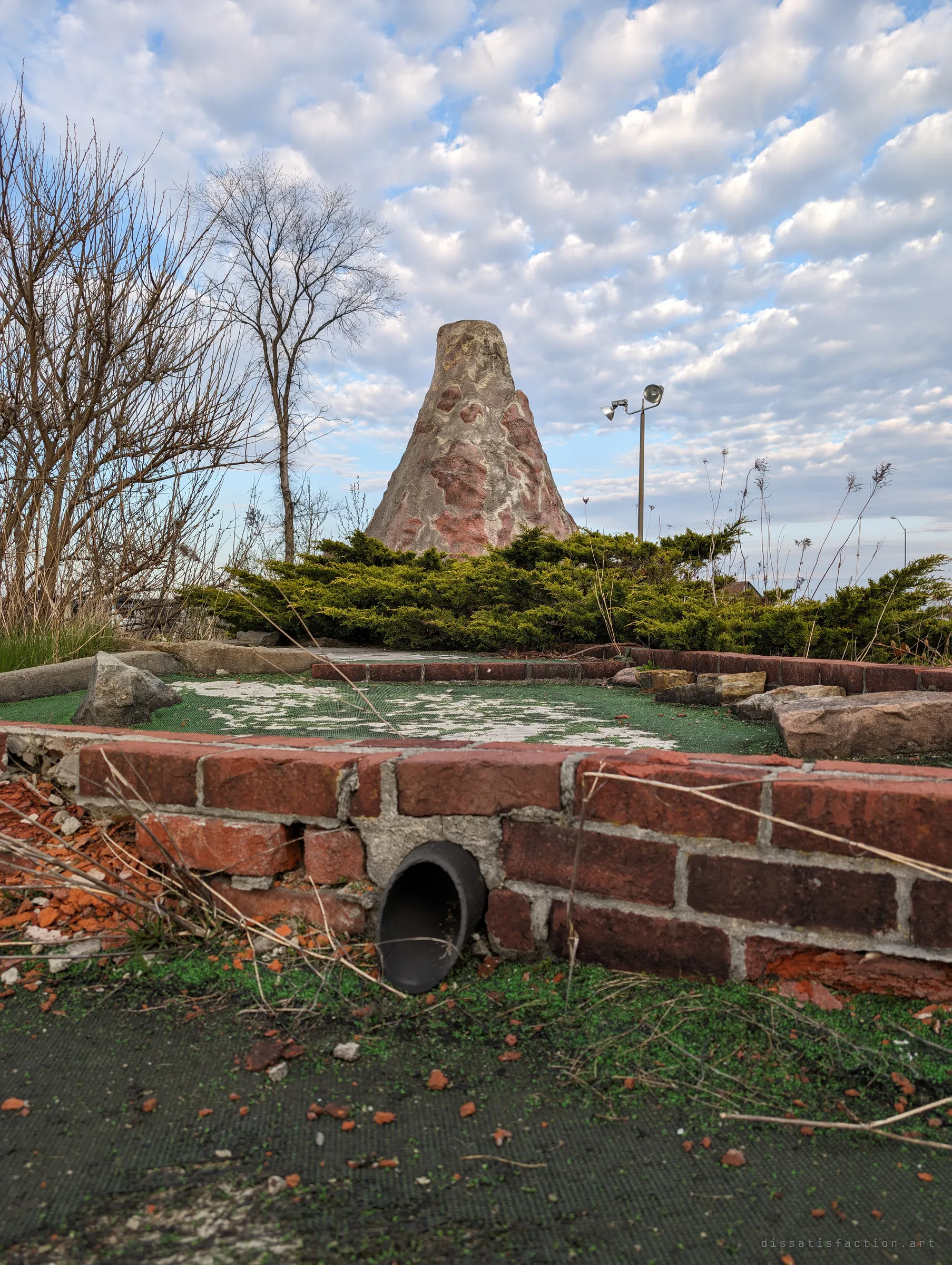 A cracked brick-edged mini golf hole with worn green carpet, a fake volcano rising behind a juniper hedge. A floodlight pole stands against a sky of puffy clouds.