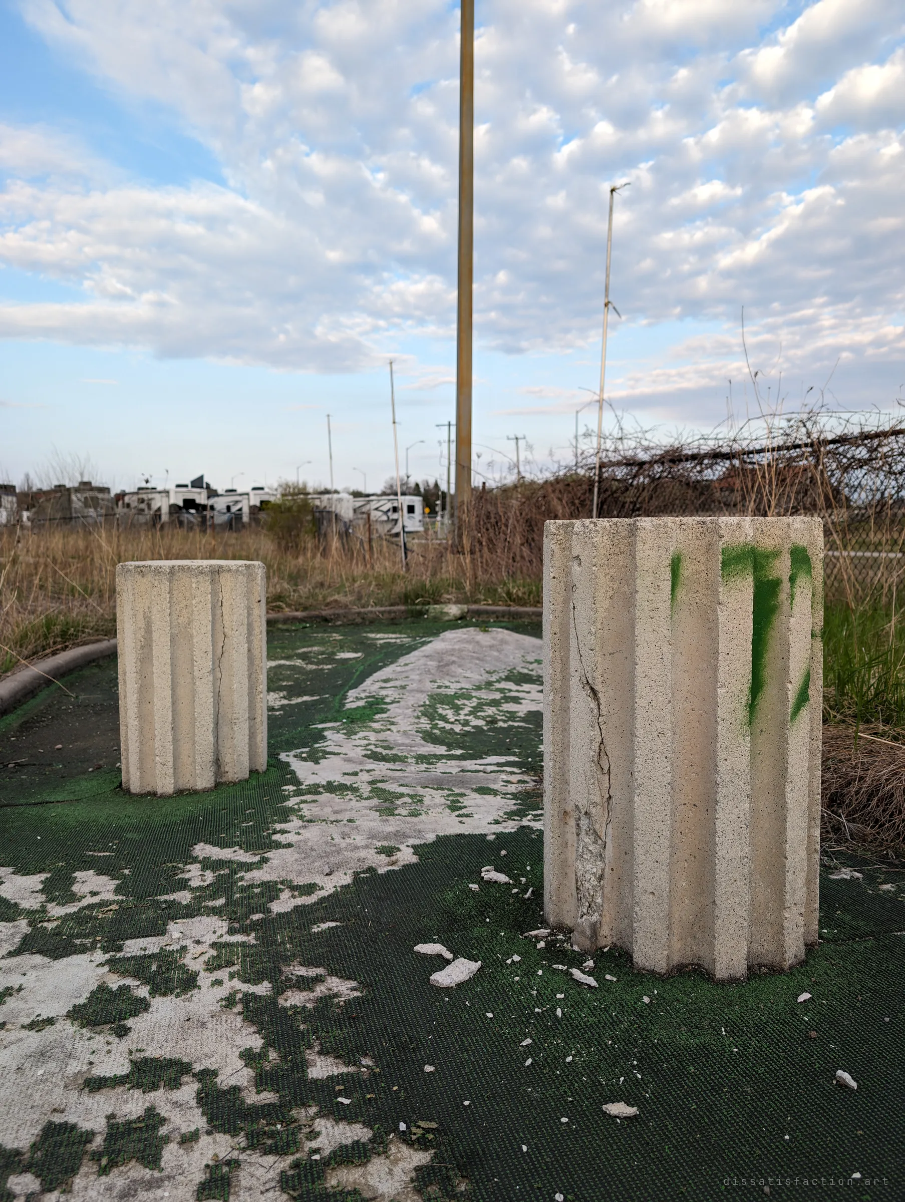 Two fluted concrete columns stand as obstacles on a mini golf green where the carpet has worn through to bare concrete. Dry grass and a chain-link fence close off the course.