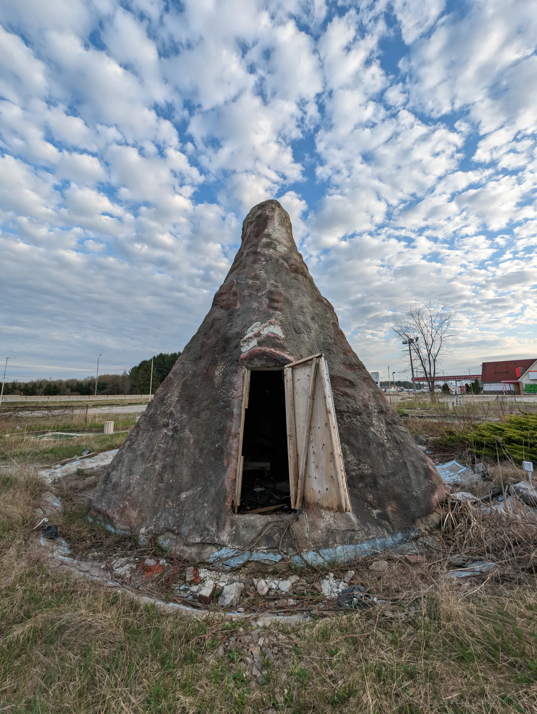 The fake volcano up close, its painted shell weathered and a makeshift plywood door hanging open into darkness. Dead grass and rubble ring its base under a dappled sky.