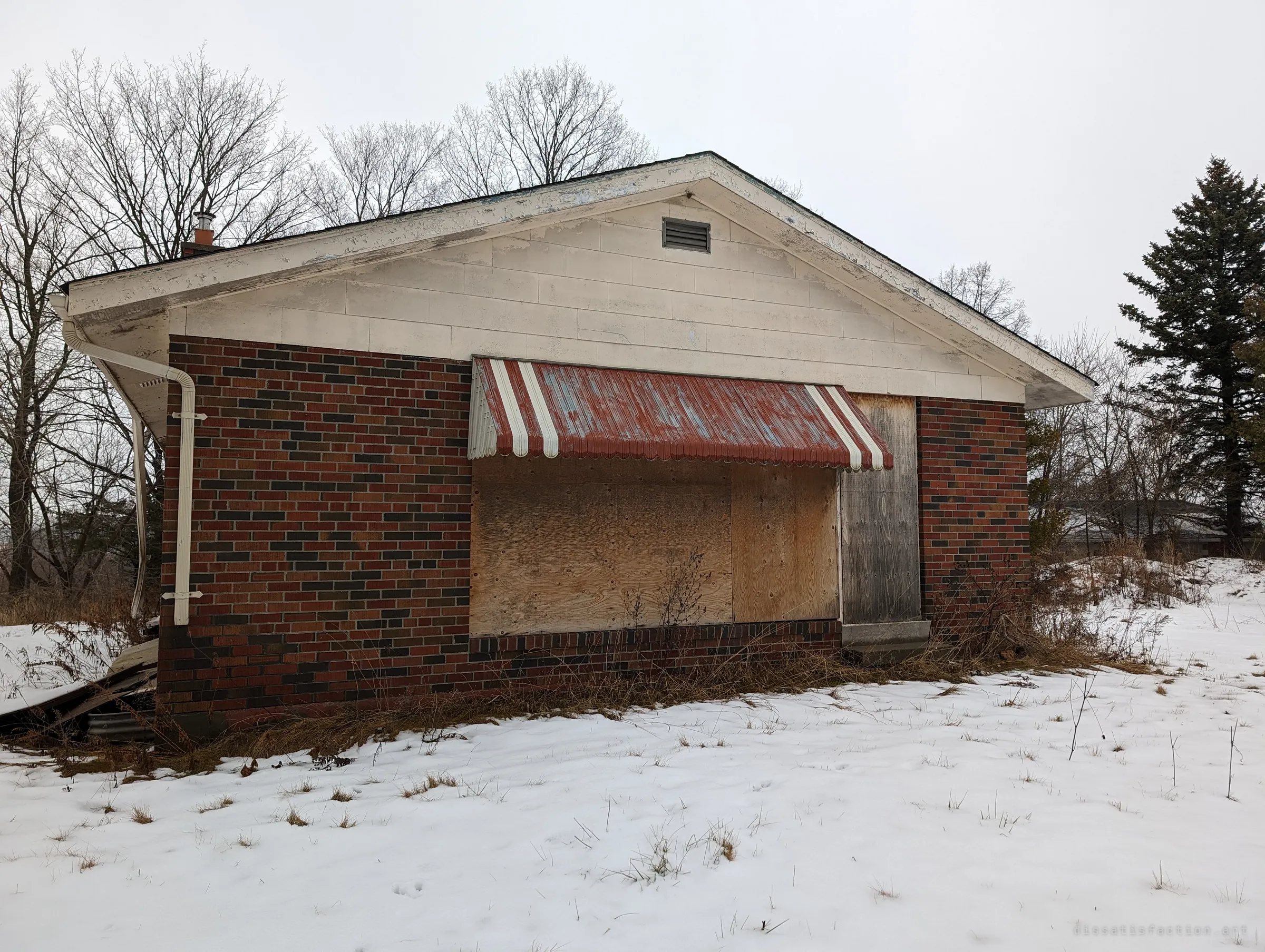 A small brick bungalow with a faded striped awning over boarded-up windows, standing in a snowy, overgrown yard. The plywood tells you the house has been shut for a while.