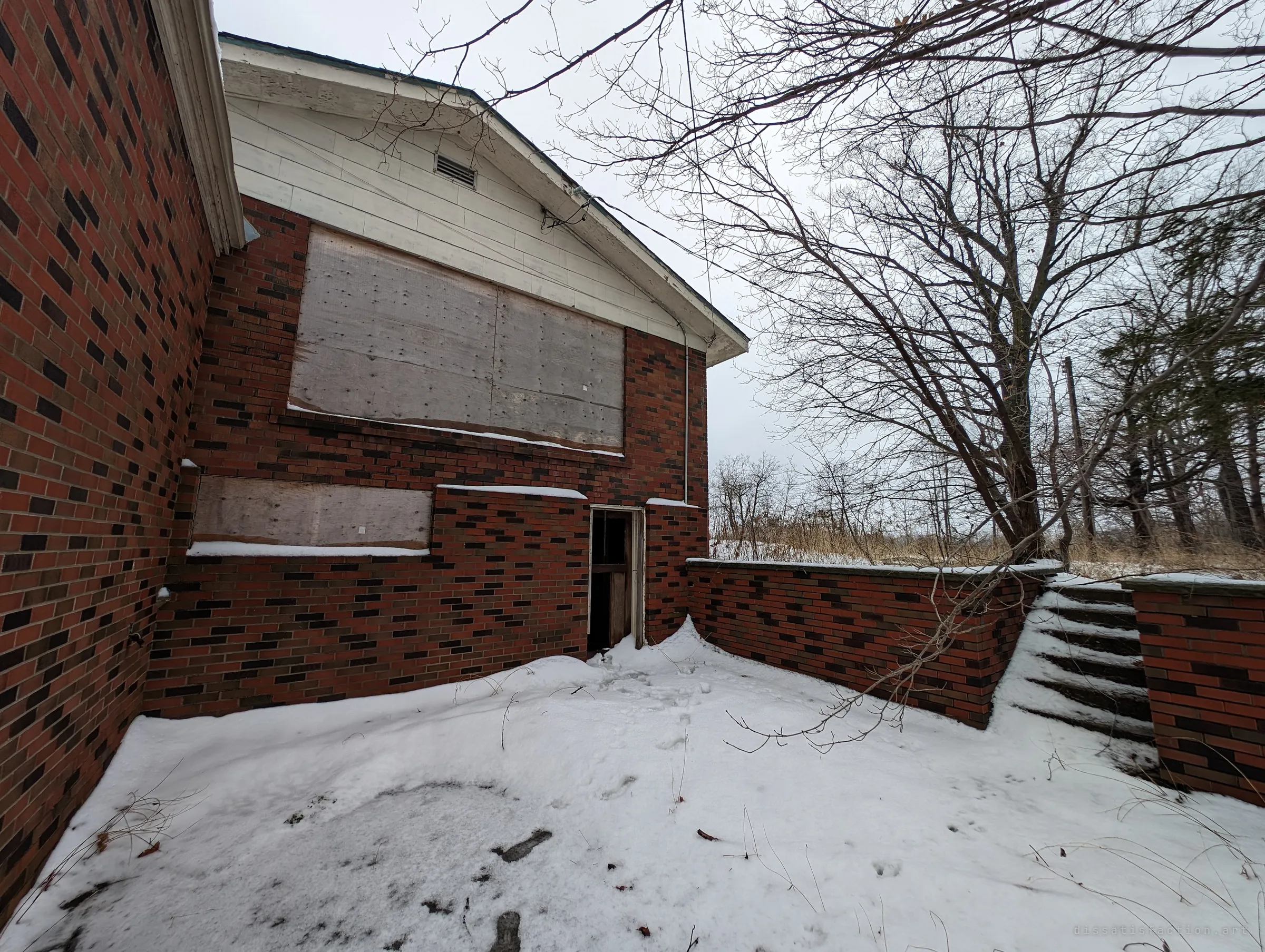 Wide view of the back of the house: boarded windows above a narrow open doorway, with a brick-walled terrace and snowy steps leading off to the right.