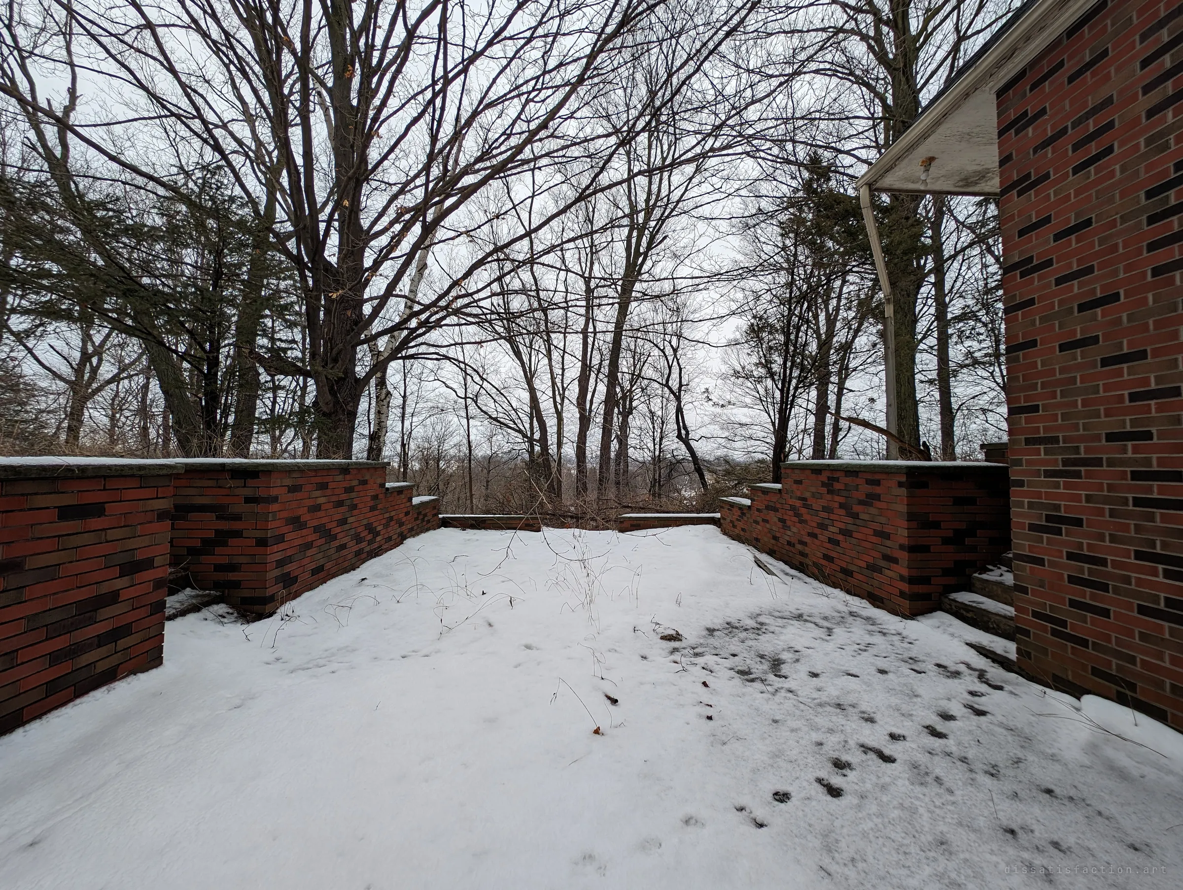A snow-covered brick terrace behind the house, its low patterned walls stepping down toward a wooded slope. Animal tracks cross the fresh snow.