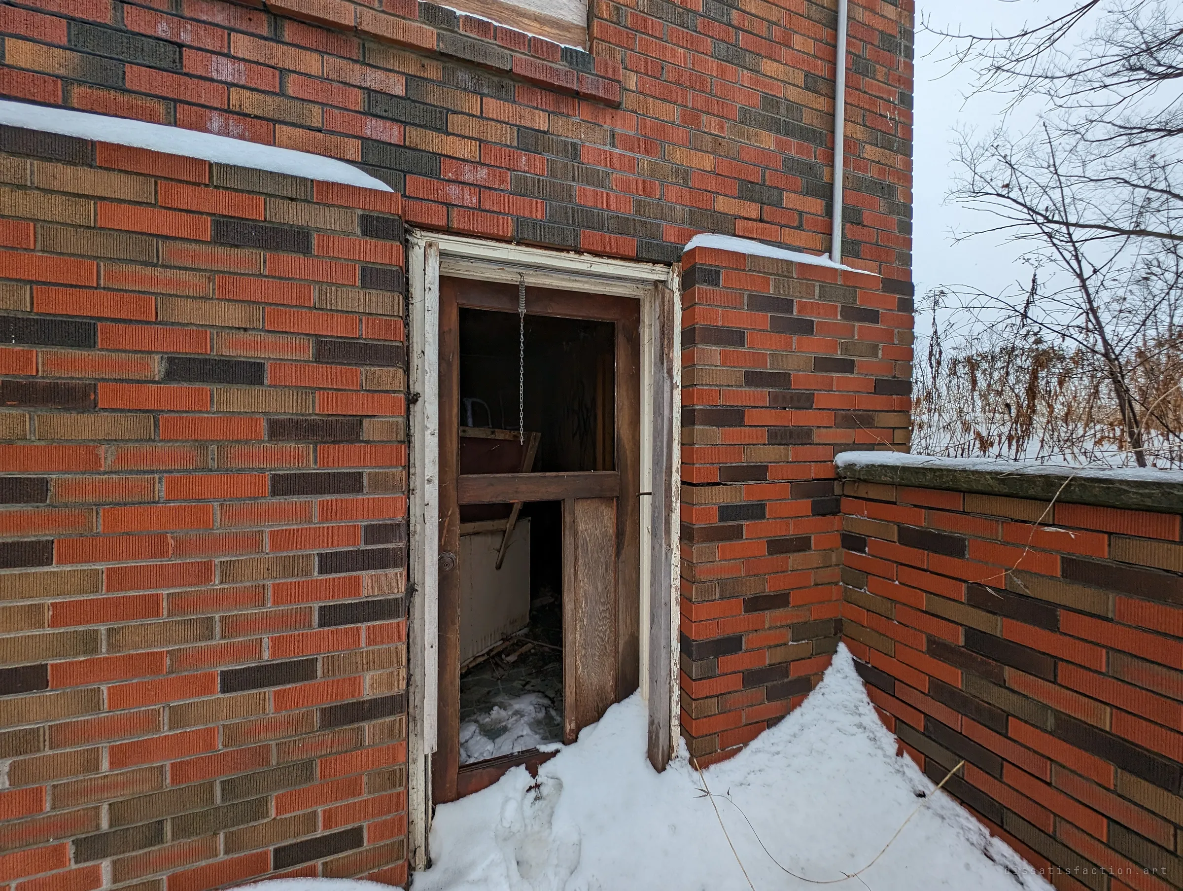 A broken wooden door hangs open in the patterned brick wall, snow drifting over the threshold. Inside, debris and an old appliance are visible in the dark.