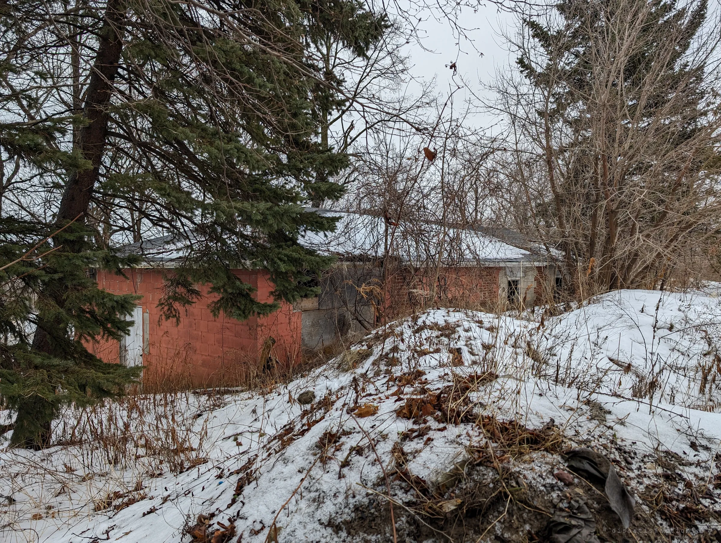 A low red outbuilding half hidden behind spruce branches and snow-covered mounds of rubble. It was left unexplored.