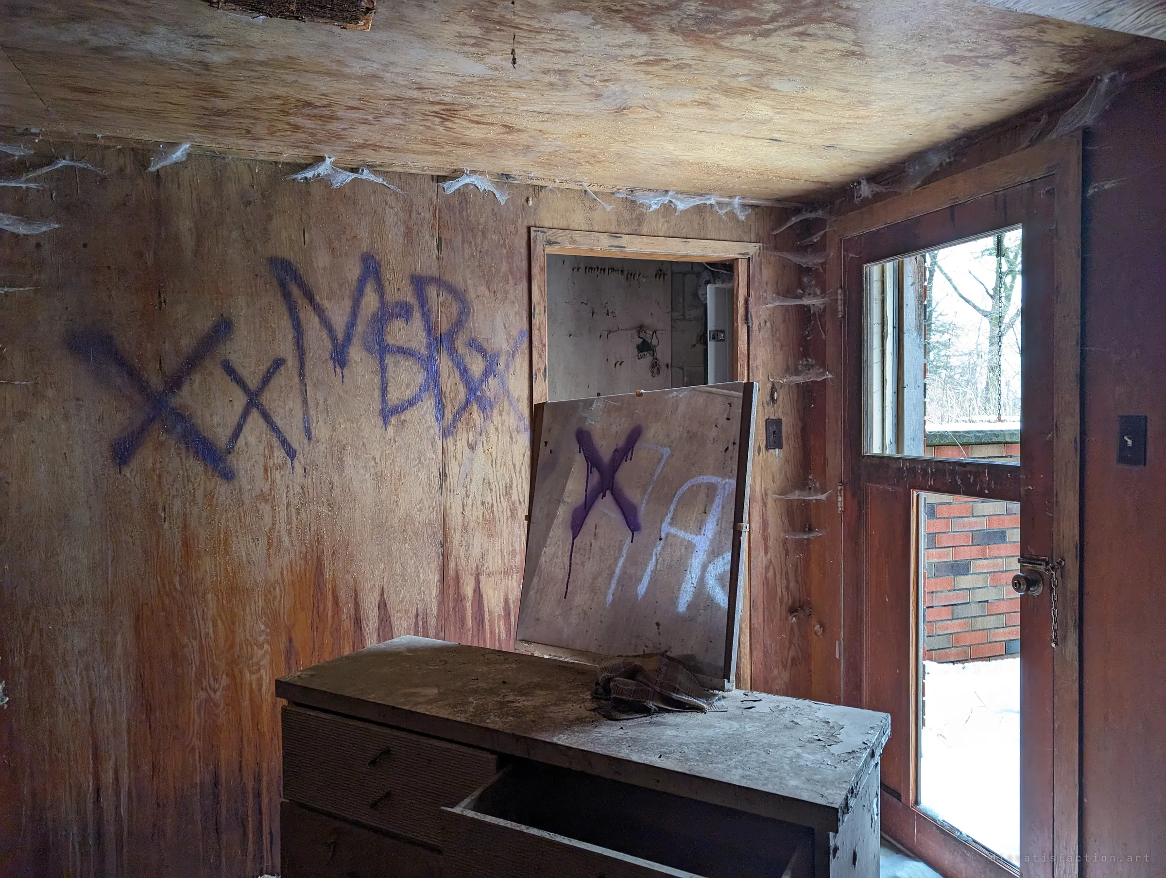 A plywood-panelled room covered in cobwebs, with purple spray-paint tags on the wall and a leaning board propped on an old dresser. Snow shows through the glass back door.