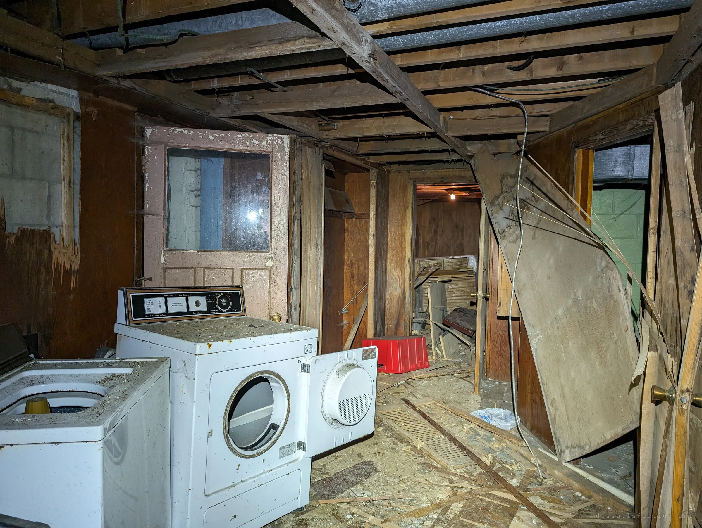 A washer and a dryer with its door hanging open sit in a gutted basement, exposed joists overhead and a smashed hallway of panelling leading back to a single lit bulb.