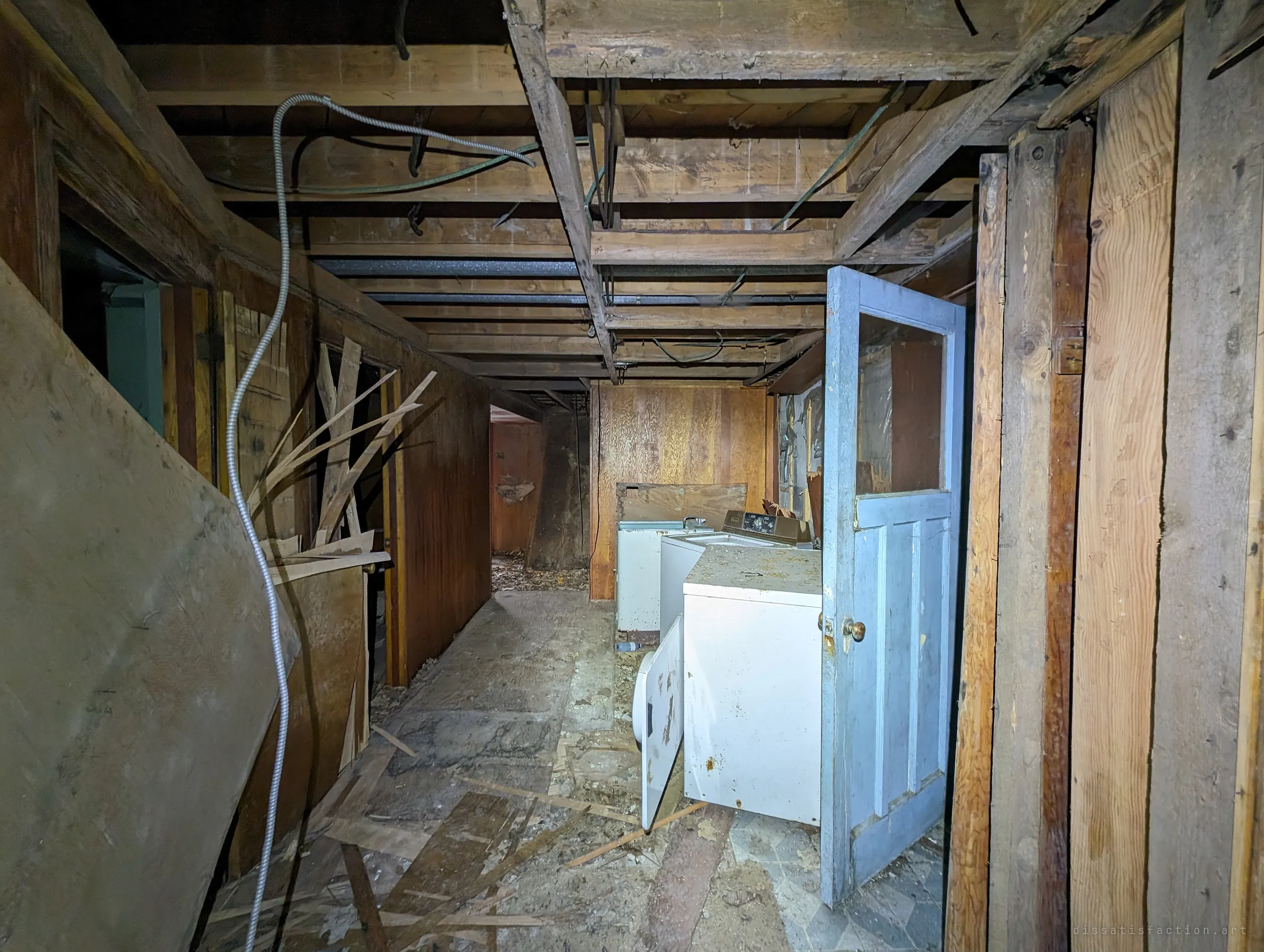 The basement corridor from the other end: loose conduit hangs from bare joists, a pale blue door stands open beside the laundry machines, and splintered panelling litters the floor.
