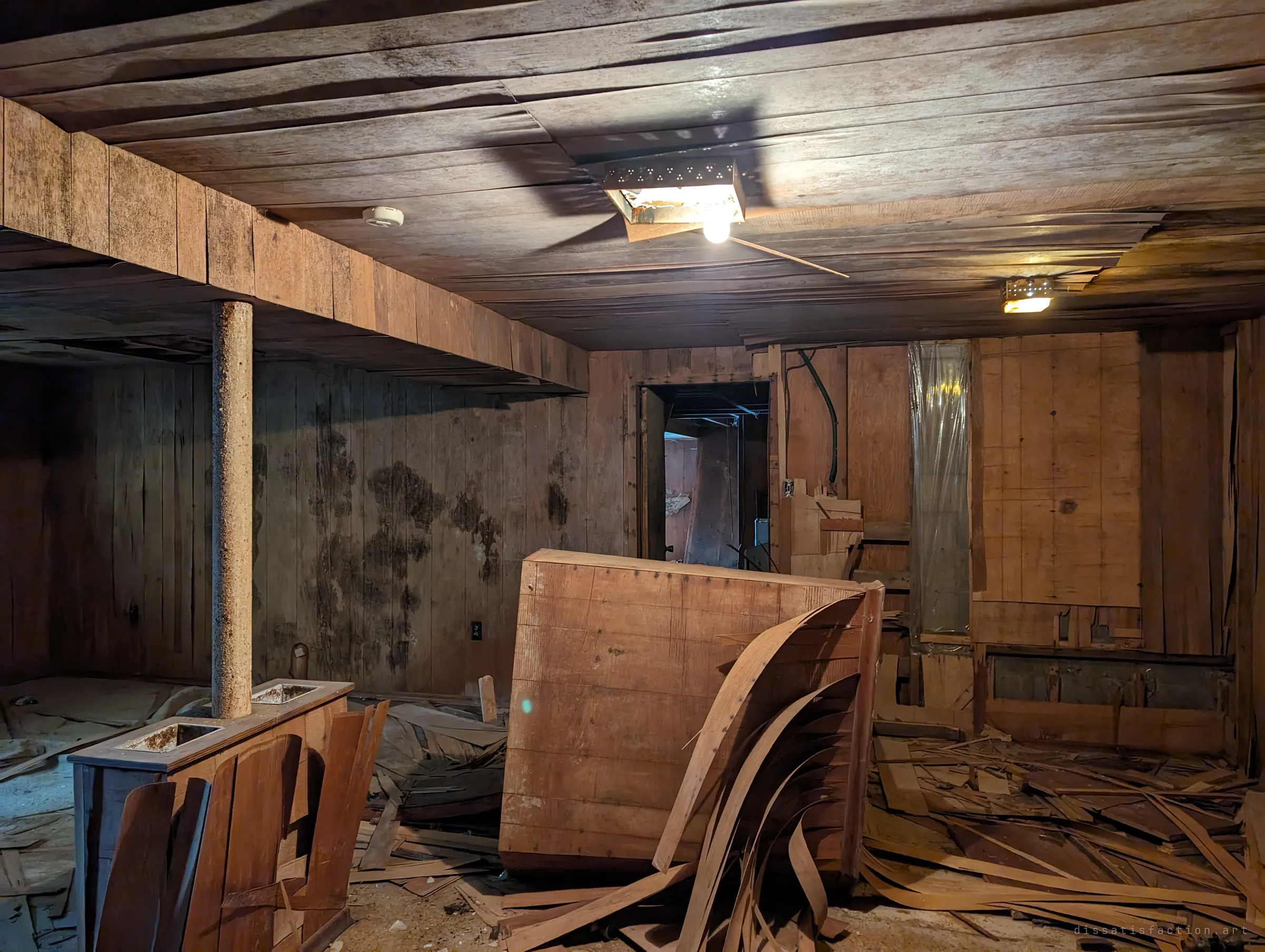 A wood-panelled basement room under a still-working light, mould spreading across the walls and curled strips of torn panelling heaped around a toppled cabinet.