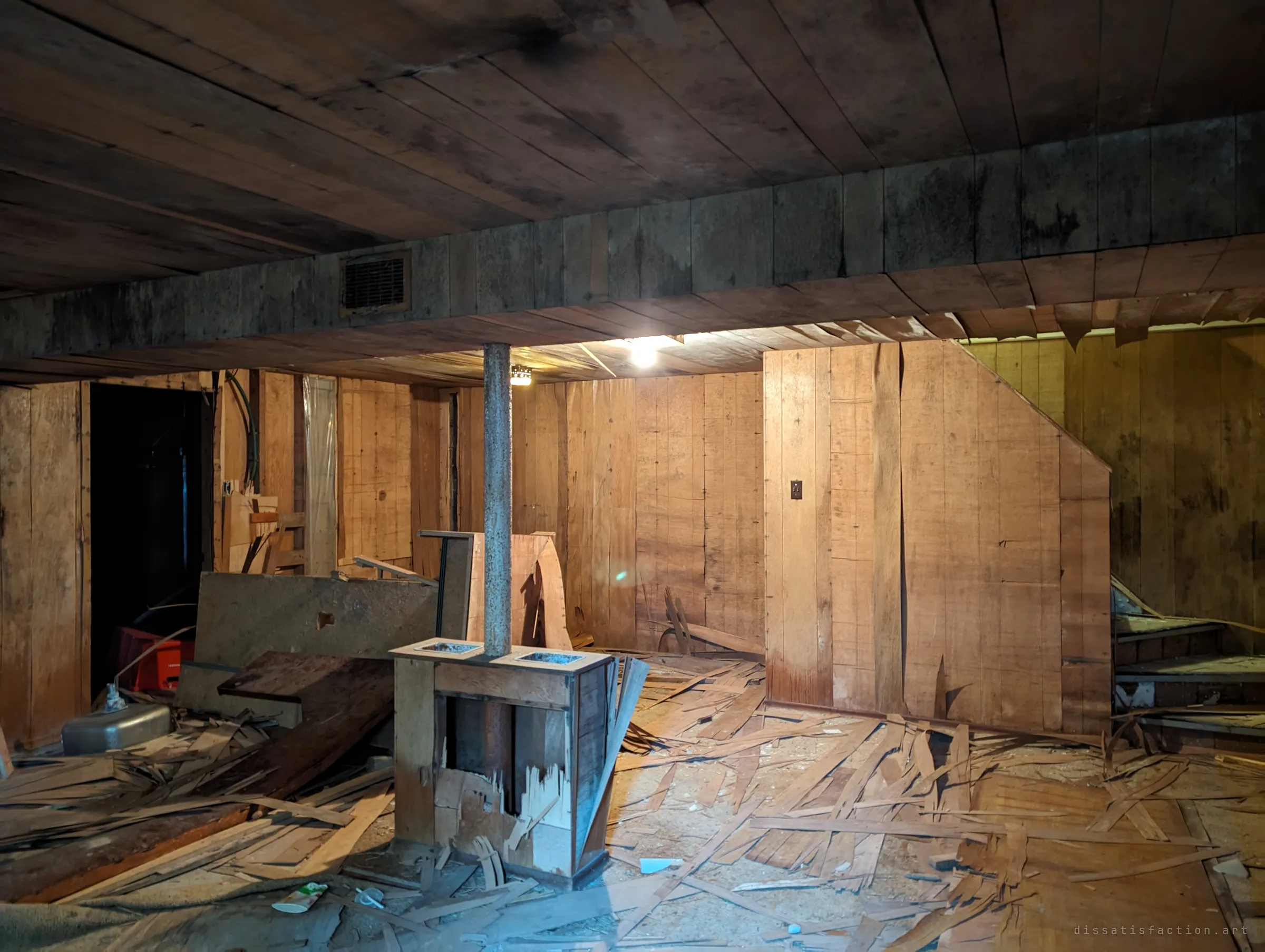 The same basement seen from another corner: a steel post rises through a broken wooden planter box, and the floor is buried in torn panelling beneath the stairs.